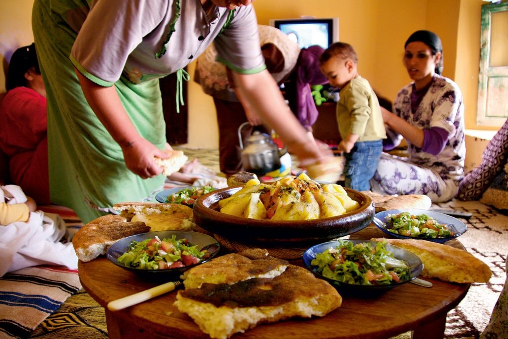 A traditional Moroccan family meal is being prepared, with a table full of dishes and people in the background, including a small child