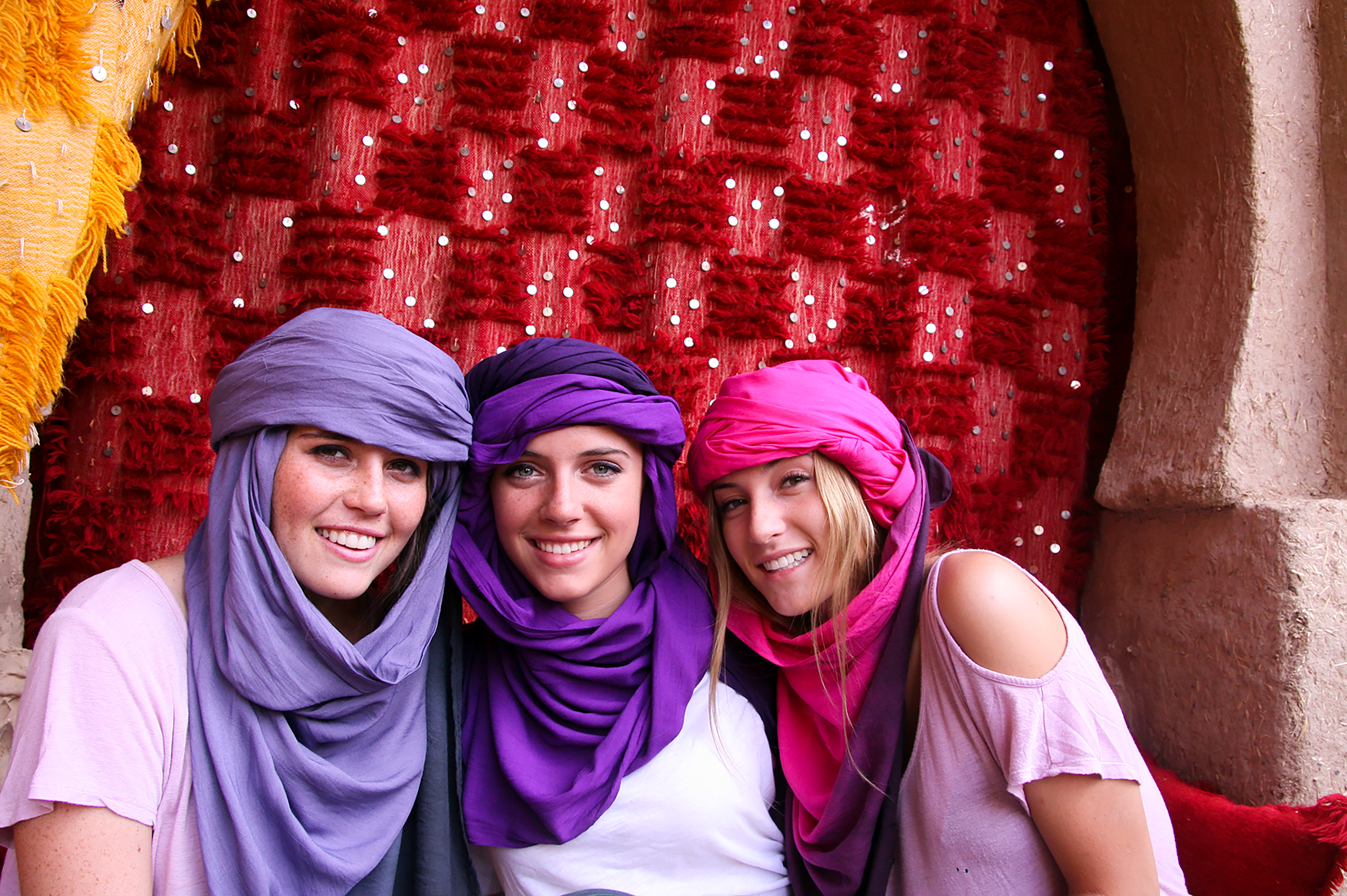 Students wear traditional headscarves in front of a patterned red textile