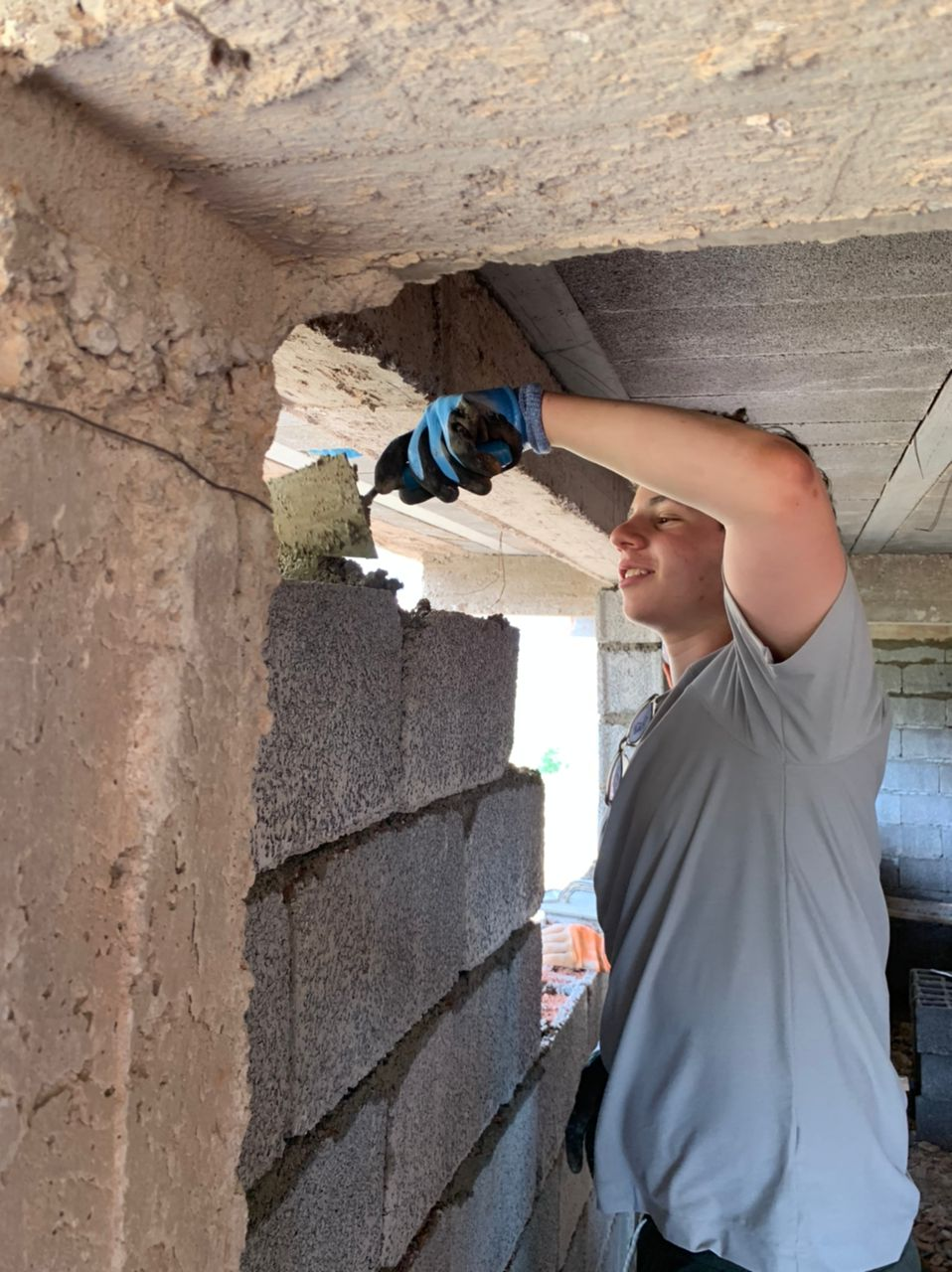 A student wearing work gloves adds mortar to a cinder block wall