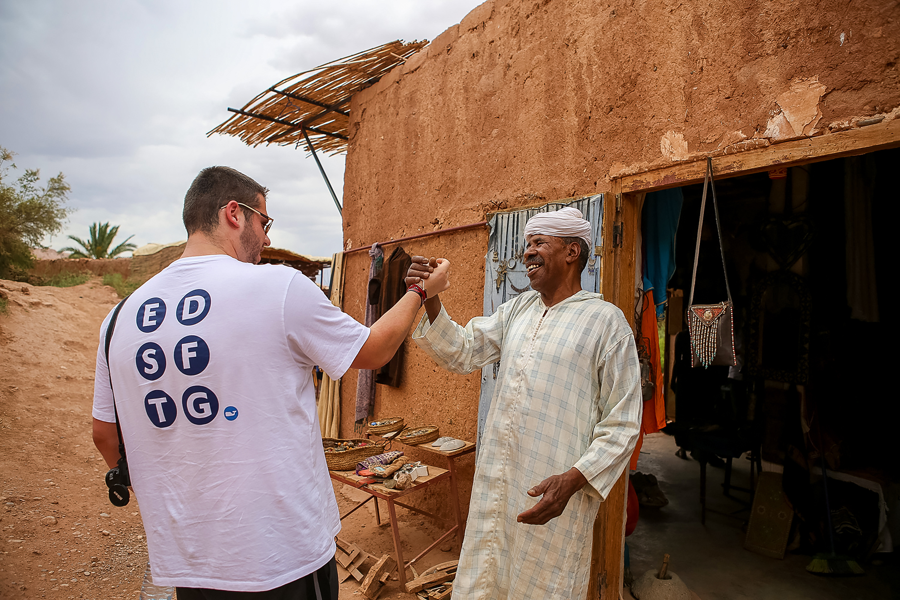 A student and a local shopkeeper share a handshake outside a mudbrick building