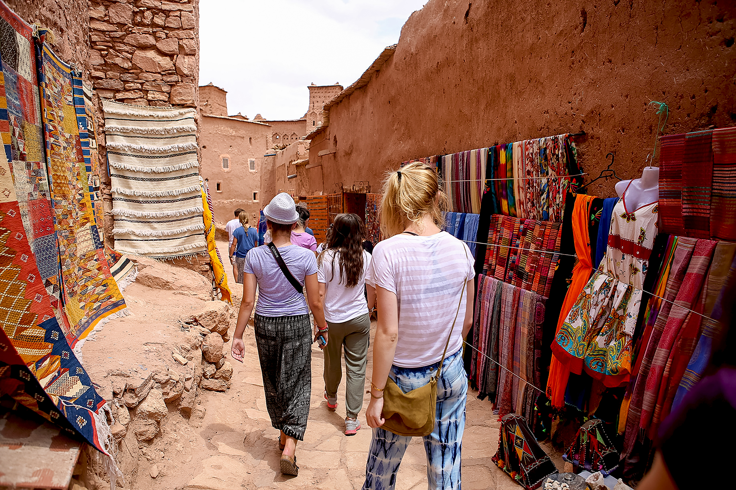 Students walk through a narrow souk alley lined with woven textiles and rugs