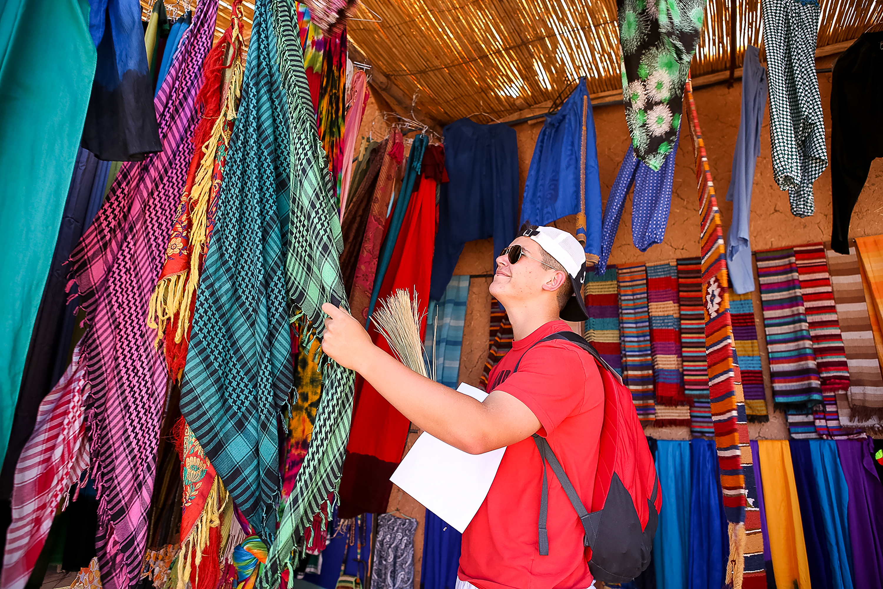 A student browses colorful woven scarves hanging in a market stall