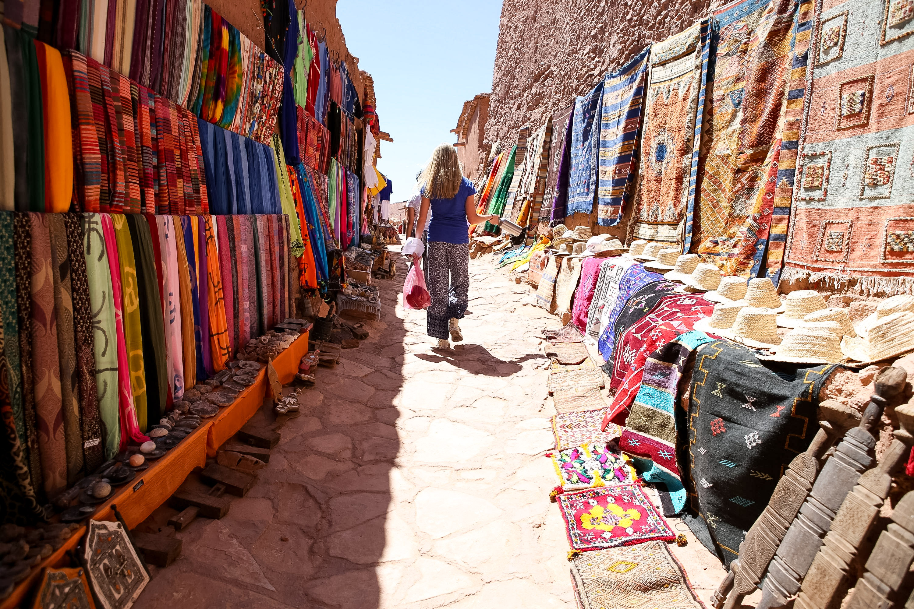 A student walks through a narrow souk lane lined with hanging textiles and woven hats