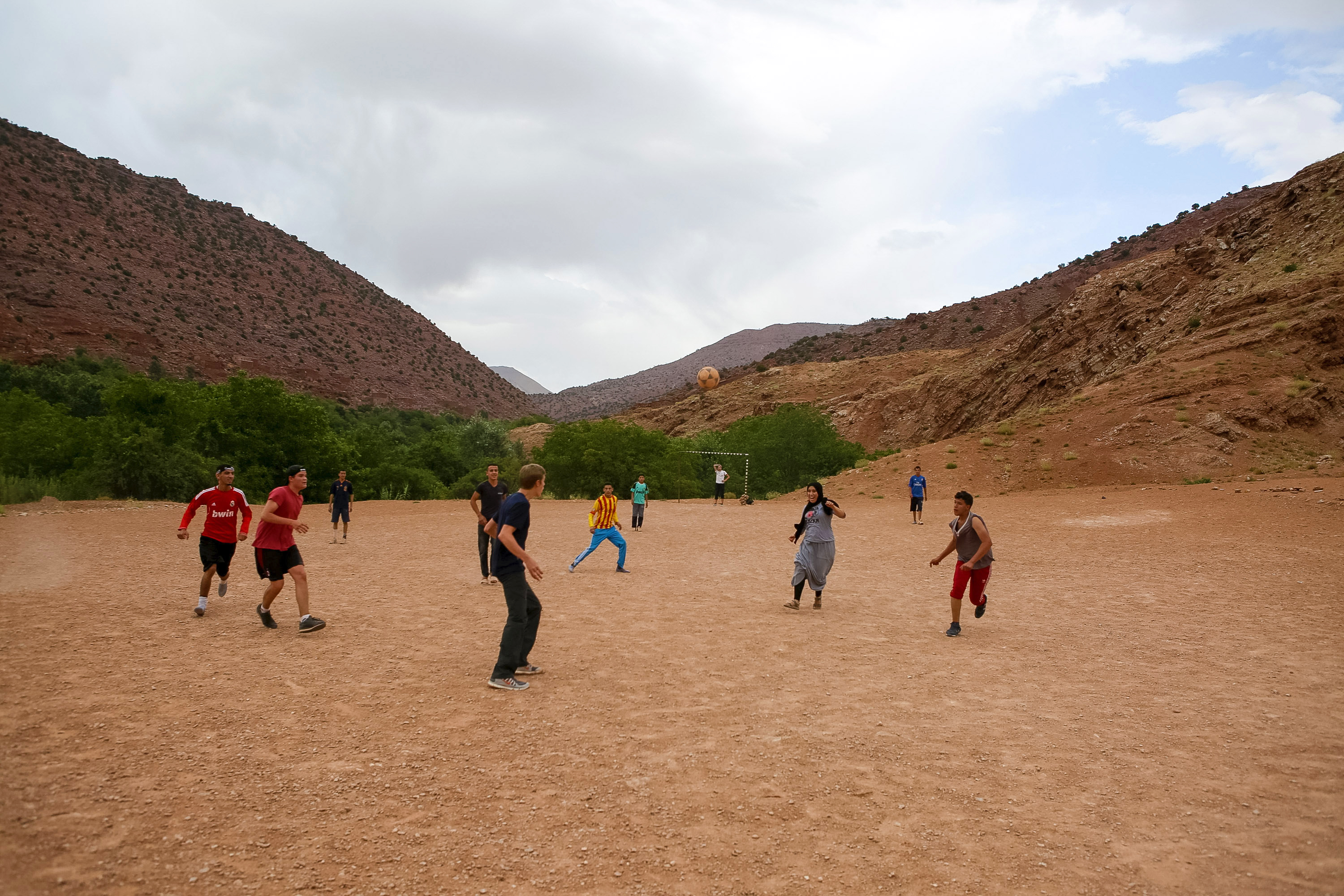Students play soccer on a dirt field surrounded by mountains