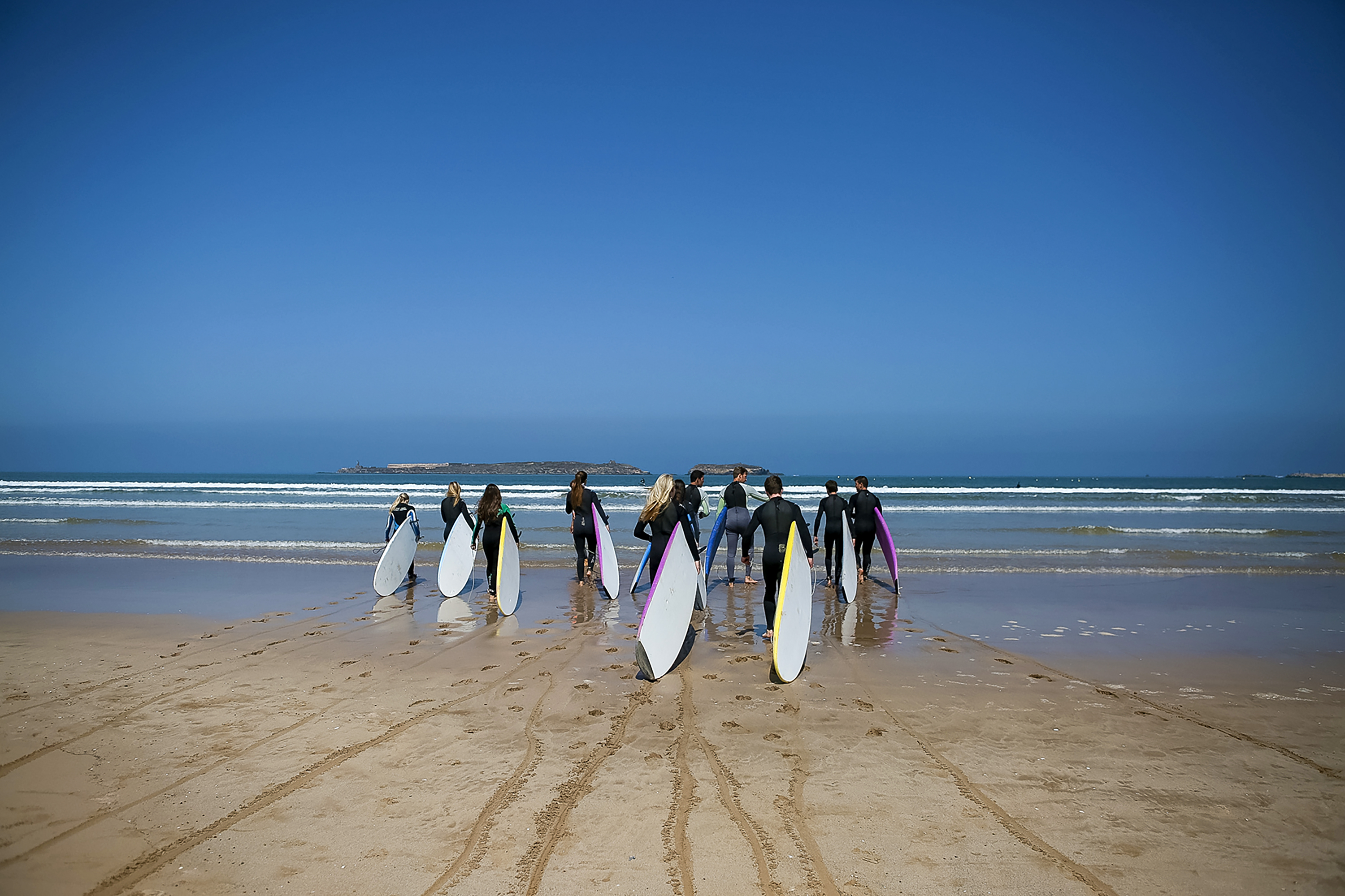Students carry surfboards across the beach toward the ocean