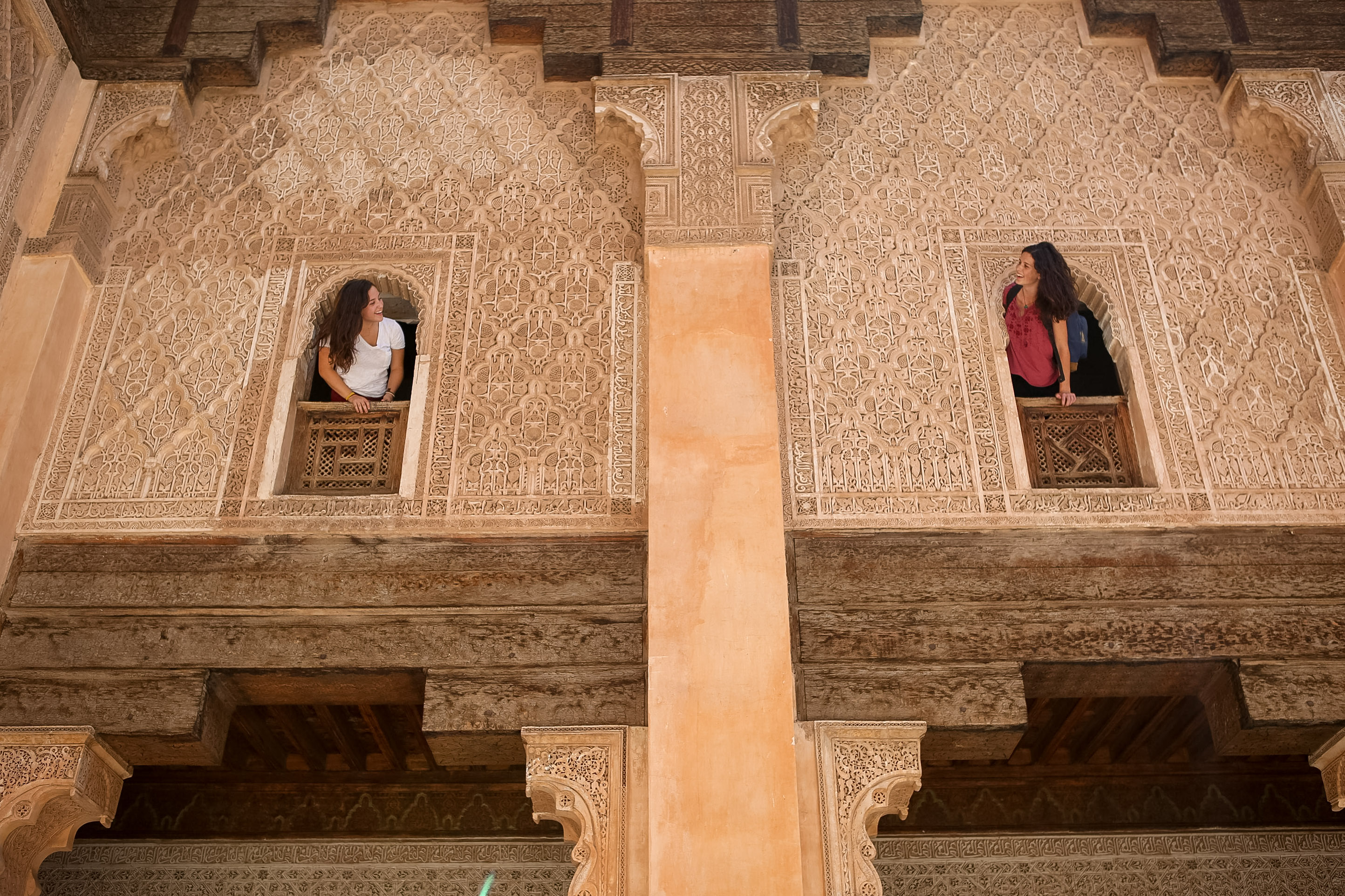 Students lean out of ornately carved window openings in a Moroccan madrasa's stone facade