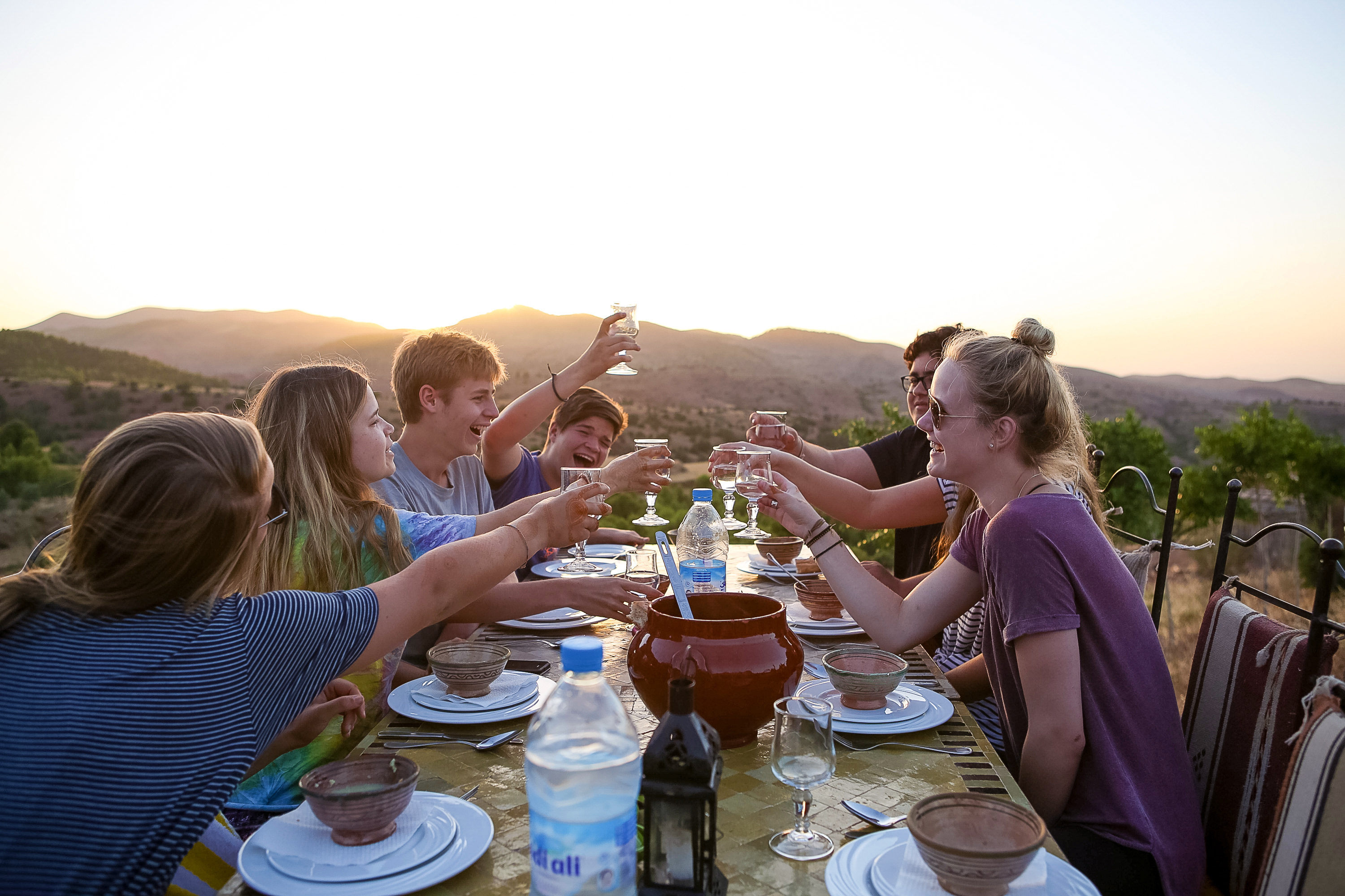 Students raise their glasses in a toast around a long outdoor dinner table set with clay bowls and a terracotta pot, with the sun setting over hills behind them.