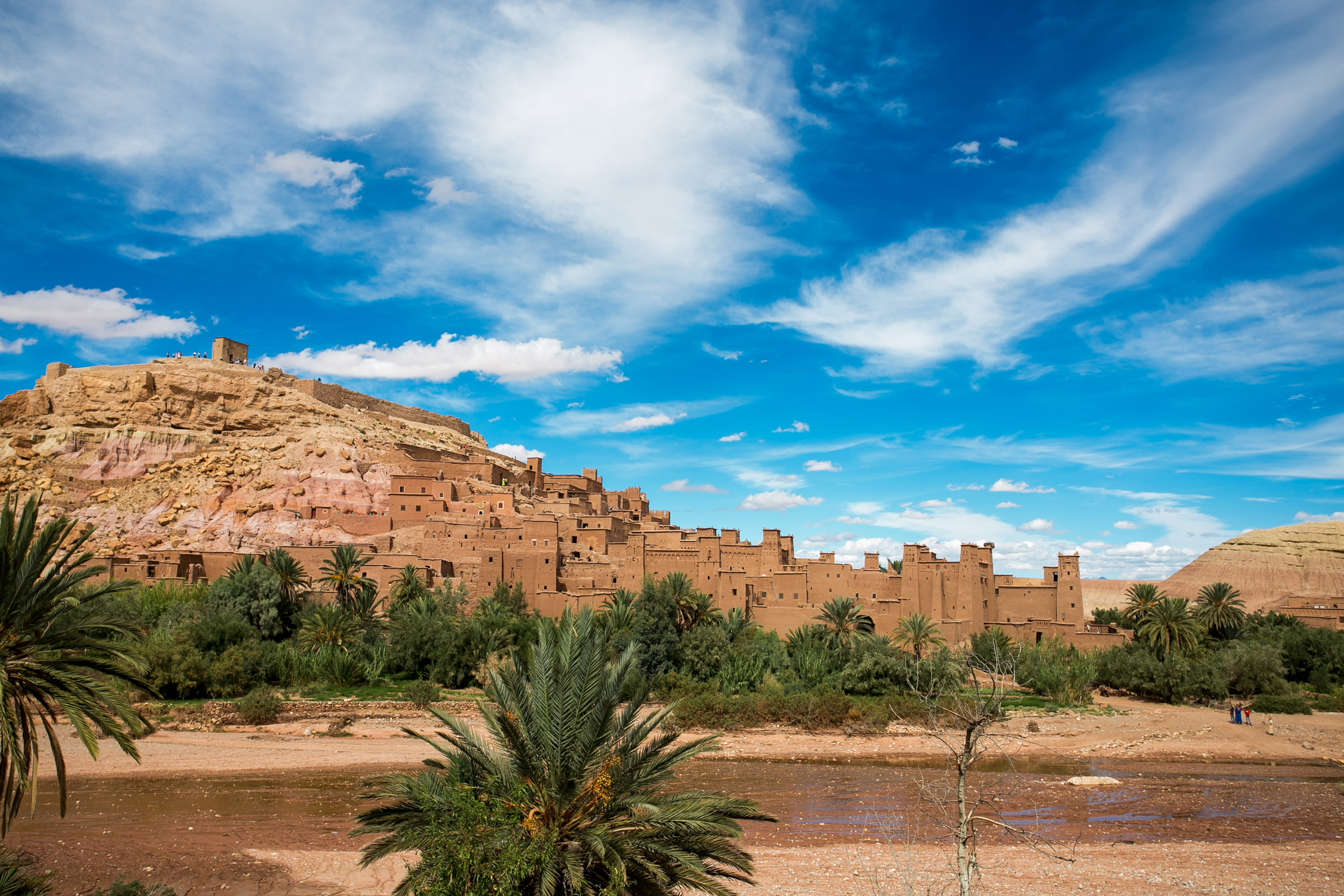 Kasbah walls rise above palm trees under a wide blue sky with wispy clouds