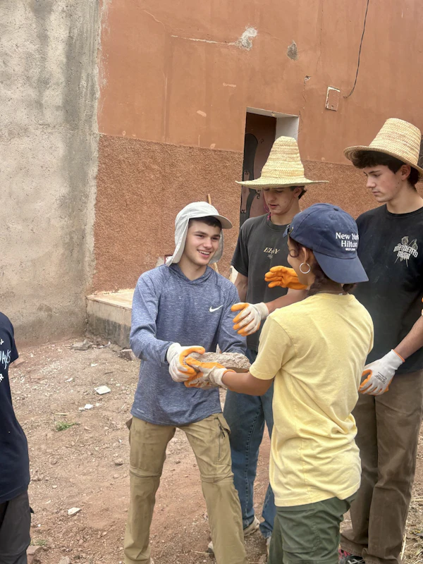 Teen travelers in work gloves and sun hats pass rocks hand to hand beside a red-earth wall on a village work site