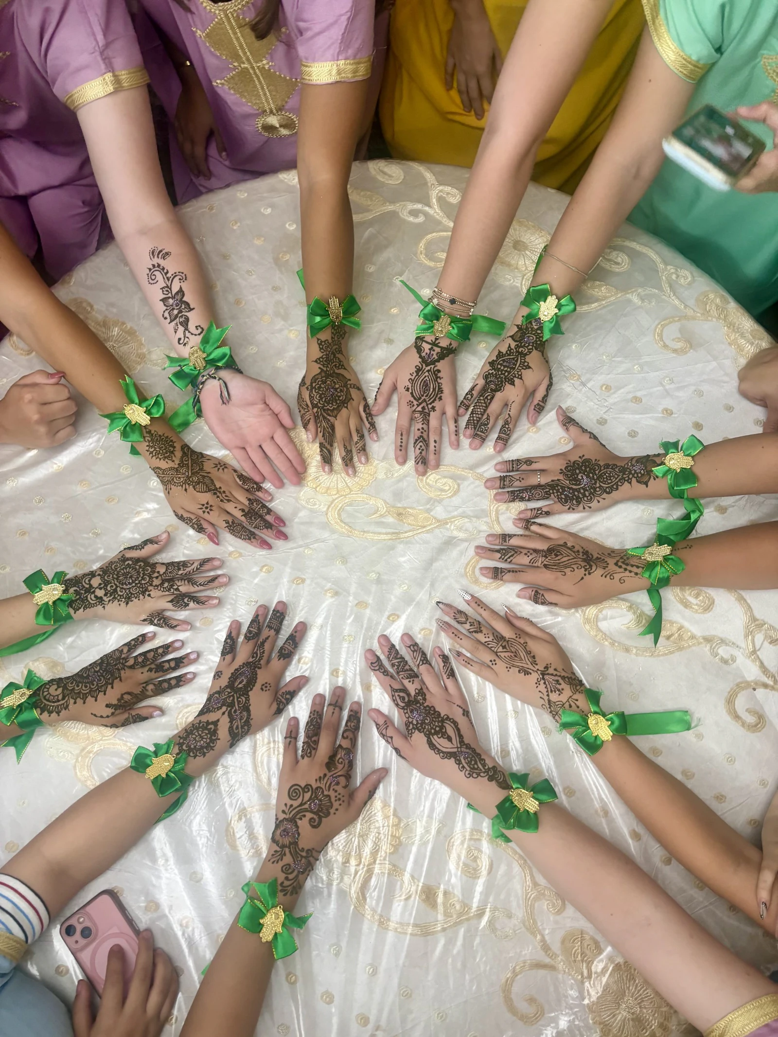 A circle of teen travelers’ hands painted with henna patterns and tied with green ribbon wristbands over a white and gold tablecloth in Morocco