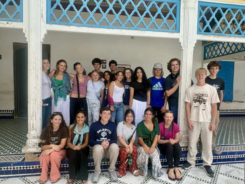 Around eighteen teen travelers stand and sit on the tiled floor of a courtyard under blue latticed balconies in Morocco