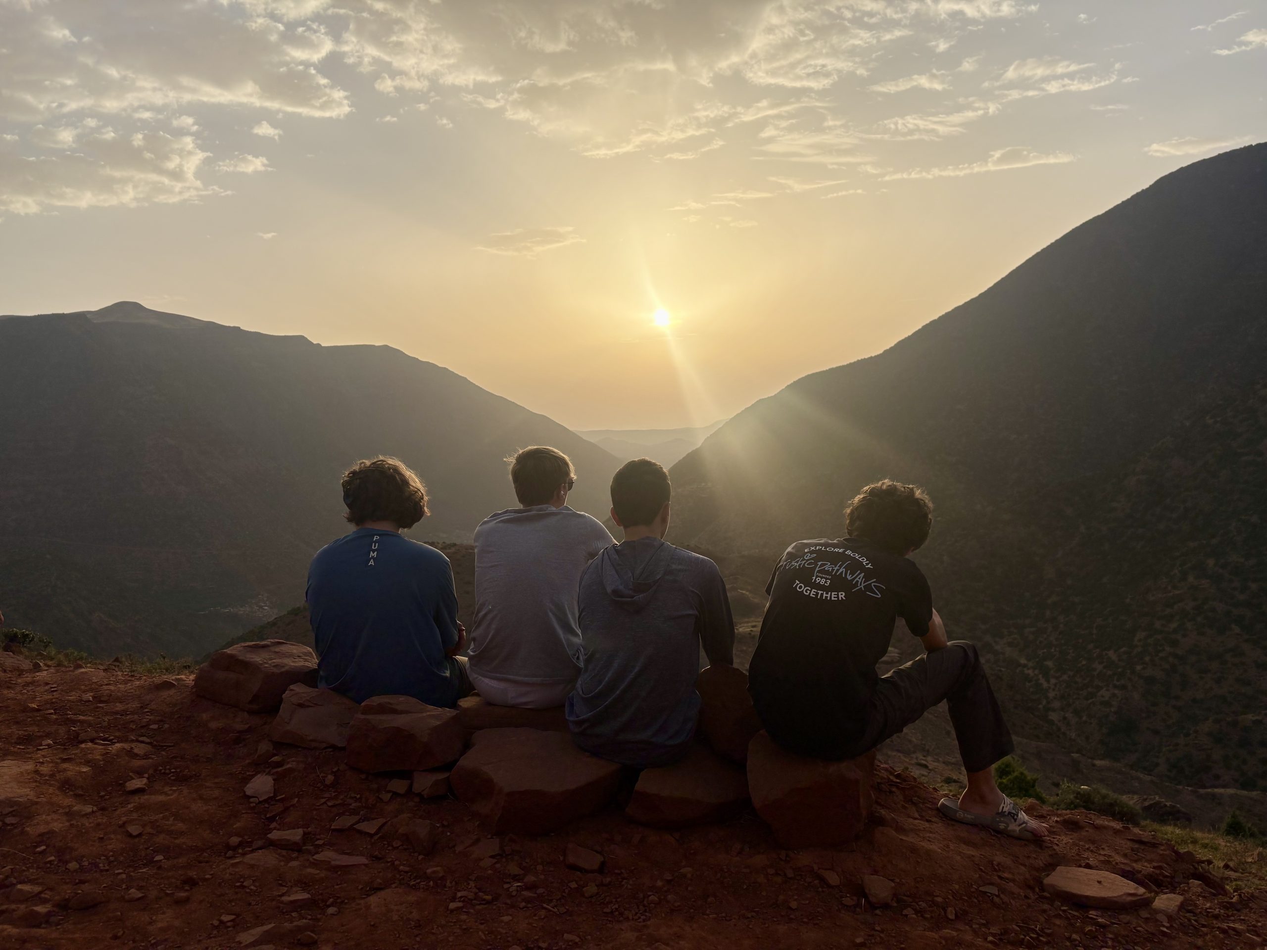 Four teen travelers sit on a rocky ledge with their backs to the camera, watching the sun set over a mountain valley