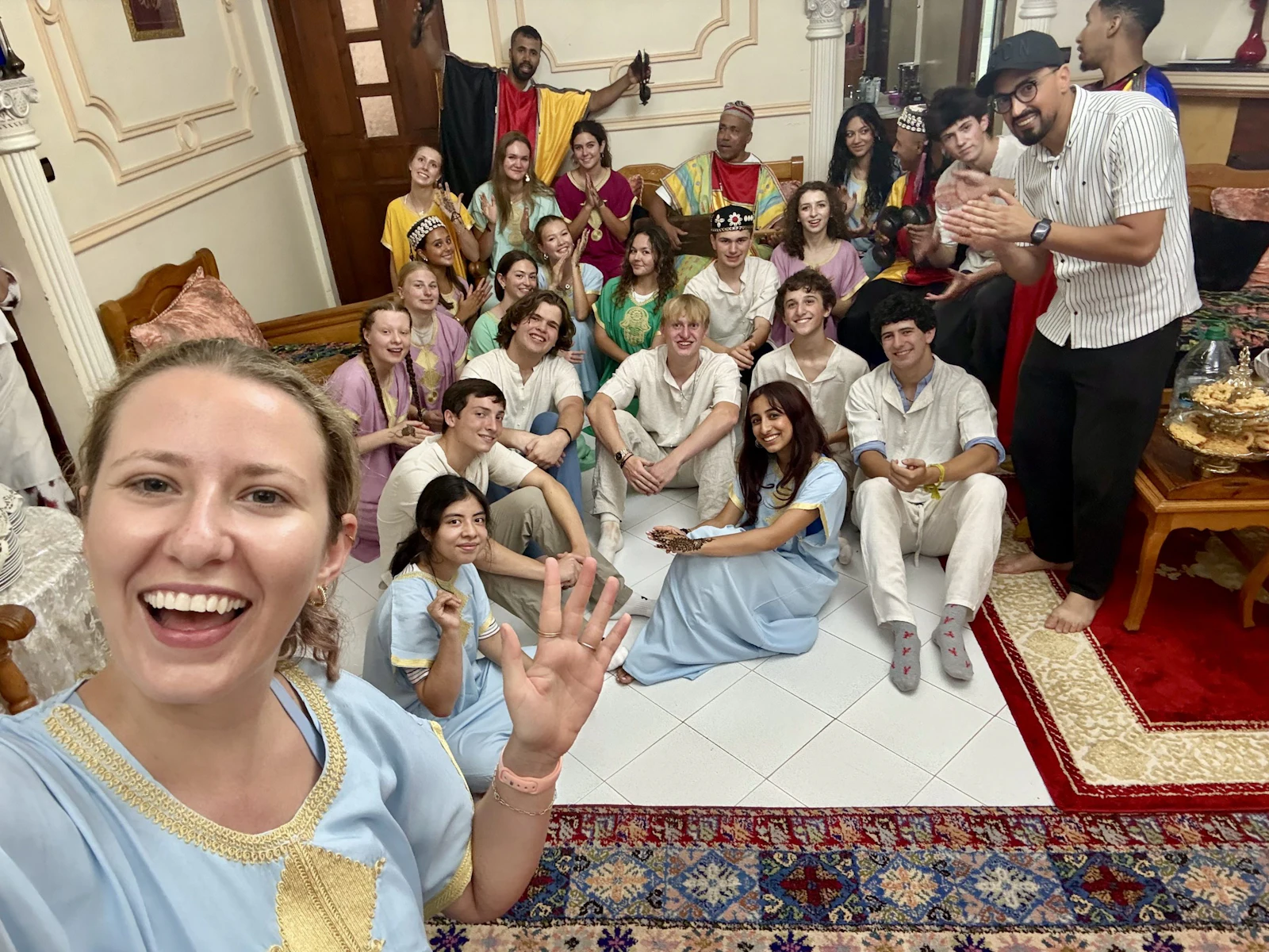 A smiling young woman takes a selfie in front of a room full of teen travelers in kaftans and djellabas on a patterned carpet