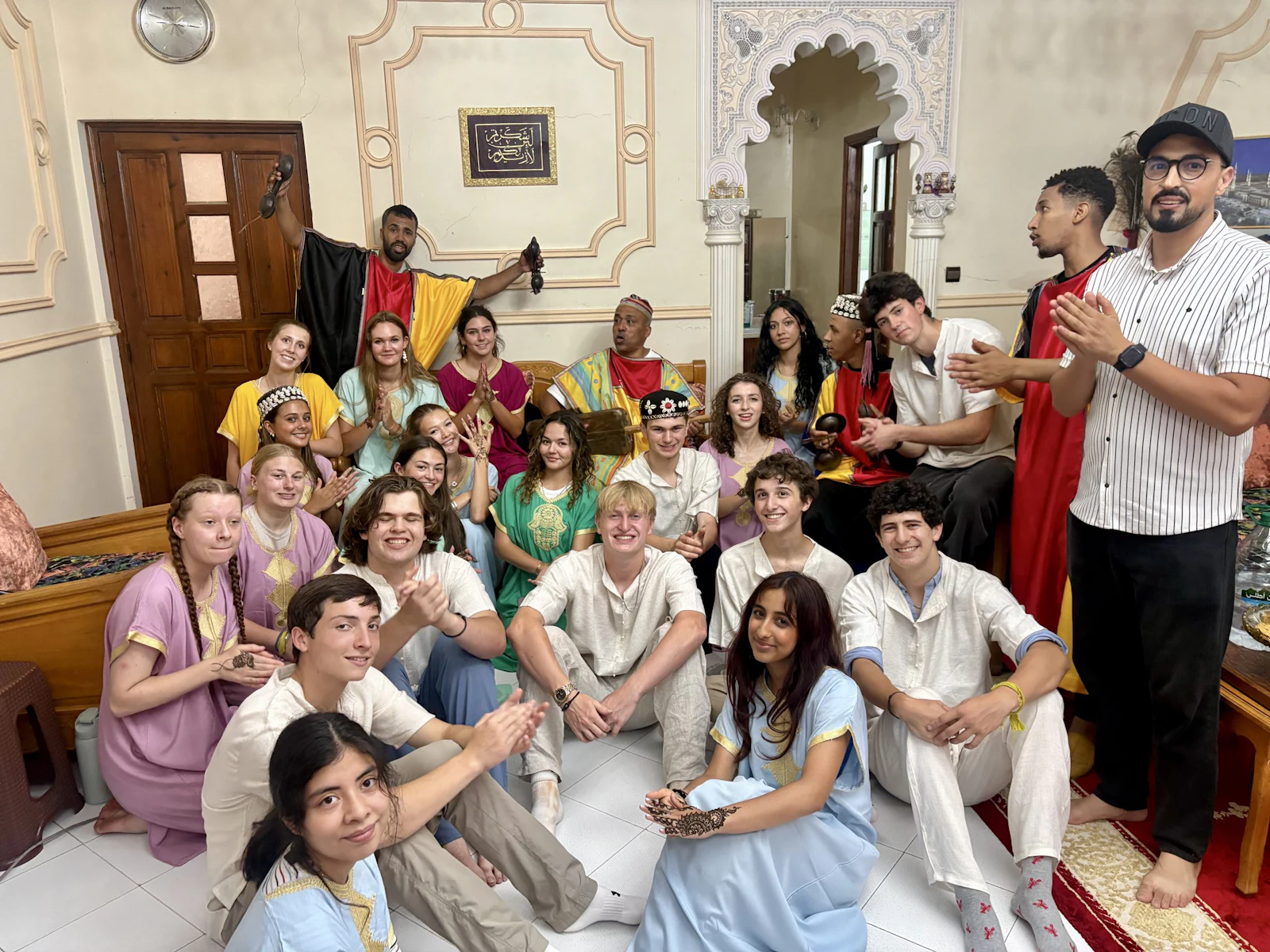 The full group of teen travelers in kaftans and djellabas sits together on the floor and banquettes of a Moroccan reception room