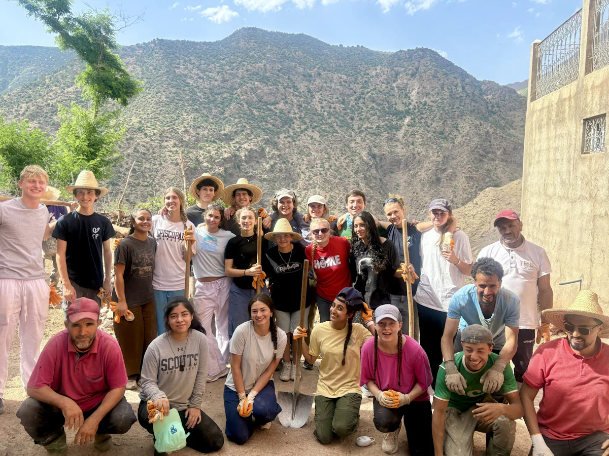 Teen travelers in sun hats and work clothes crowd together with Moroccan workers on a mountain work site, steep rocky peaks behind them