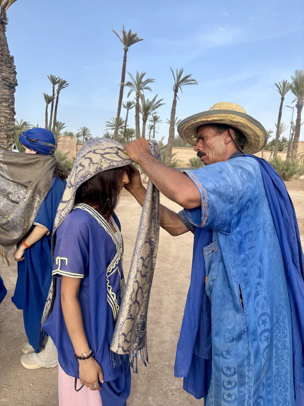 A host in a blue robe and straw hat wraps a patterned scarf around a teen traveler's head beside palm trees