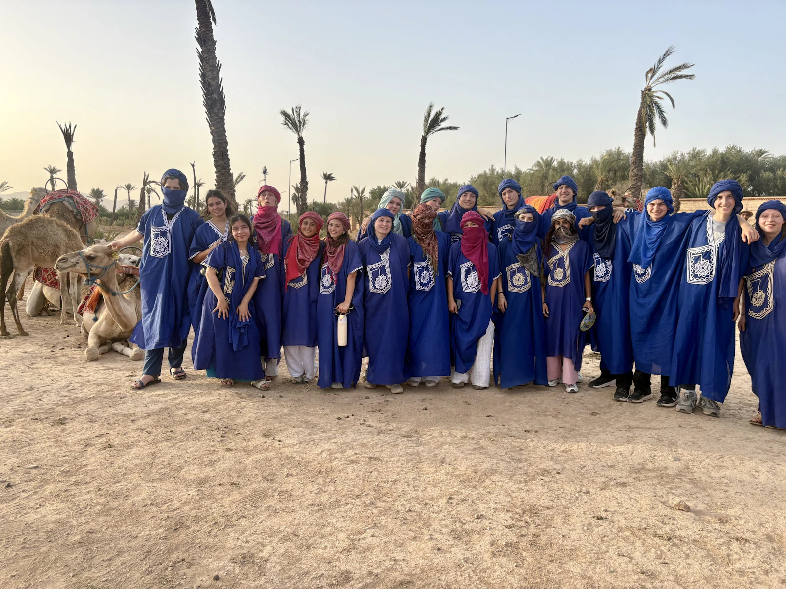 The full group of teen travelers in blue robes and headscarves stands in a long line on sandy ground beside palm trees in Morocco