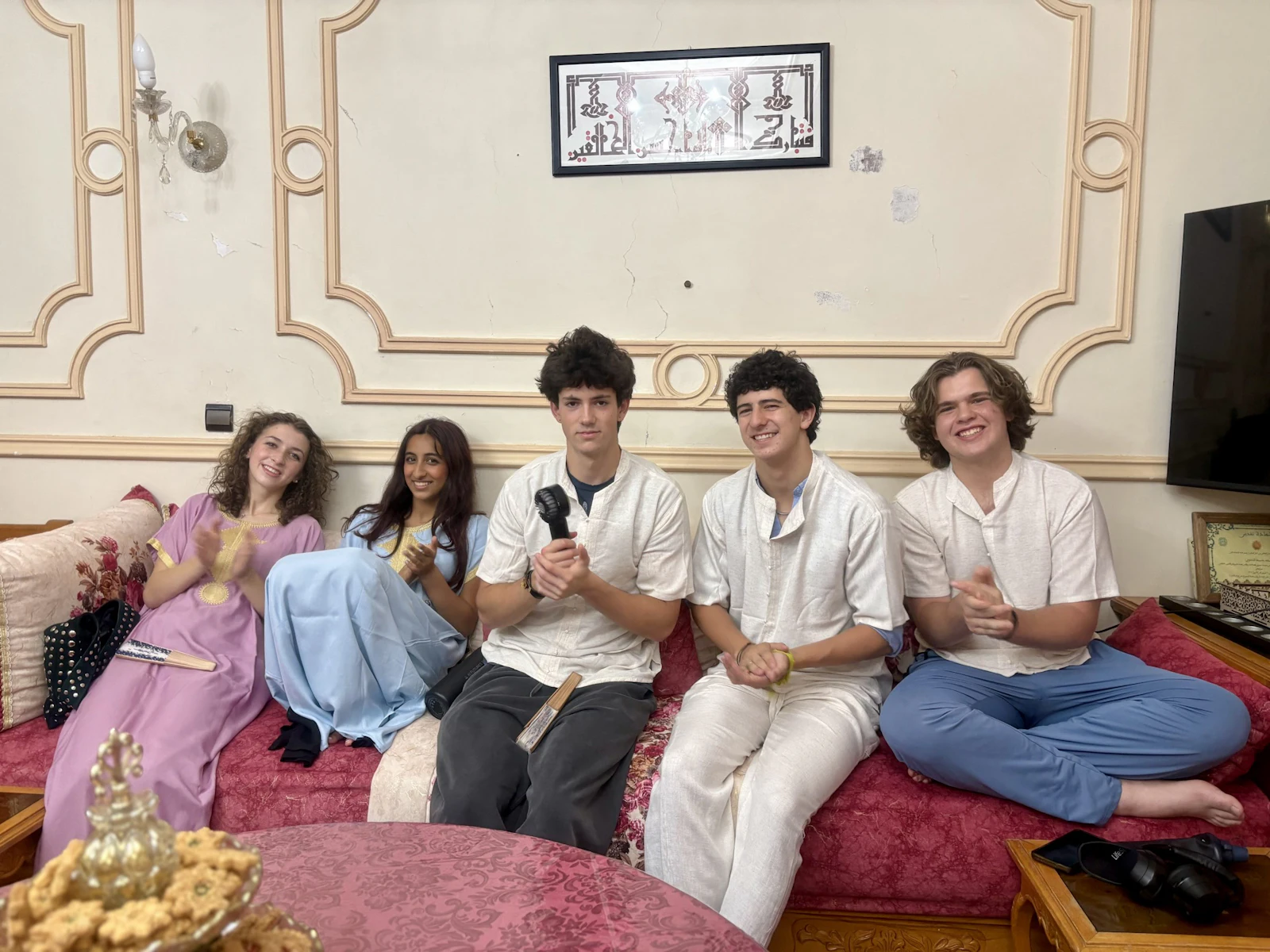 Five teen travelers in kaftans and white shirts sit along a patterned banquette in a Moroccan sitting room