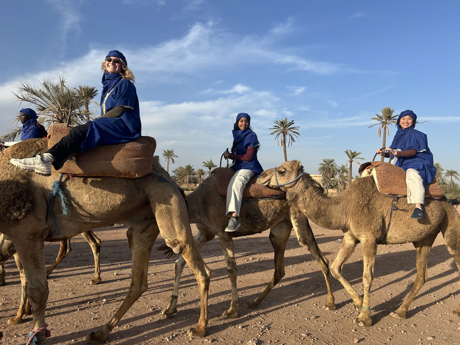 Teen travelers in blue robes and headscarves ride camels in a line past palm trees under a blue sky