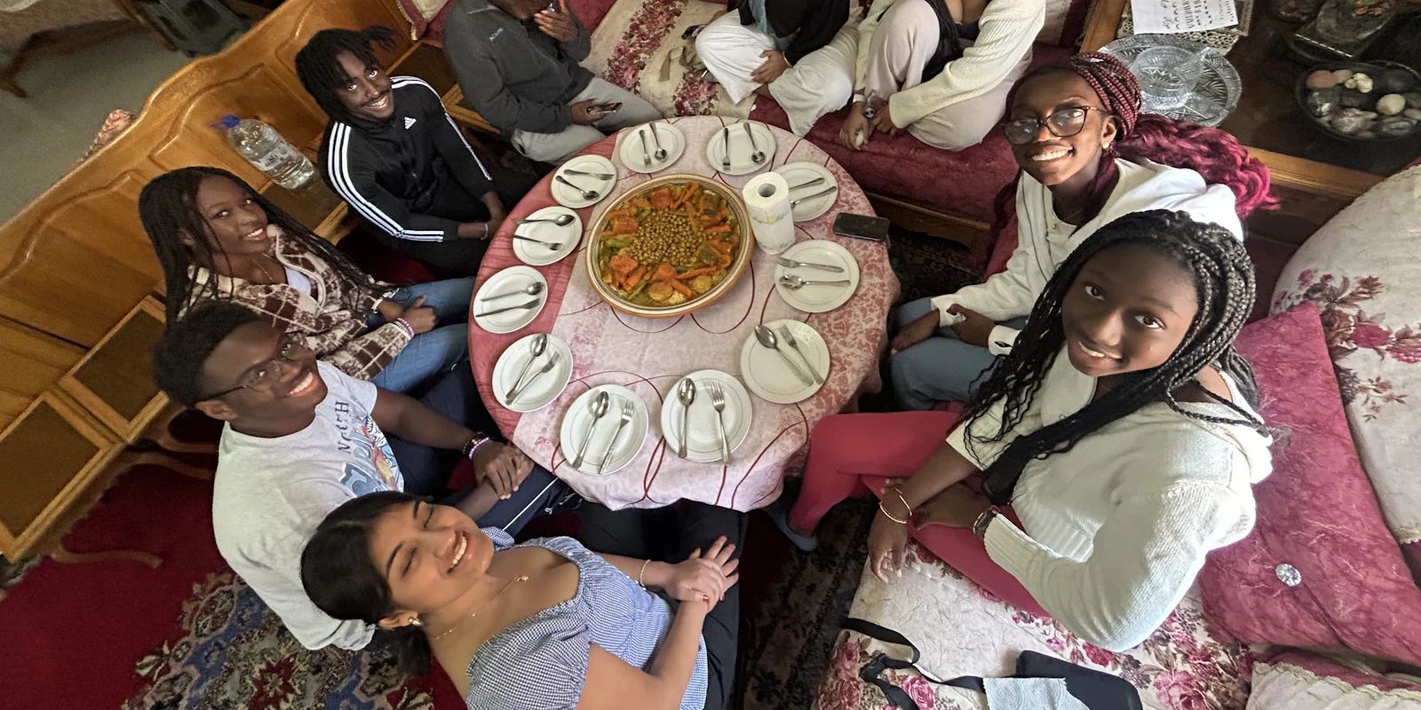 Teen travelers sit on cushions around a low table set with a large shared dish, seen from above