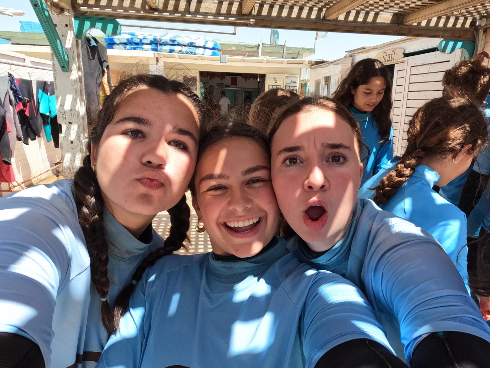 Three teen travelers in blue tops pull faces for a close-up selfie under a shaded terrace