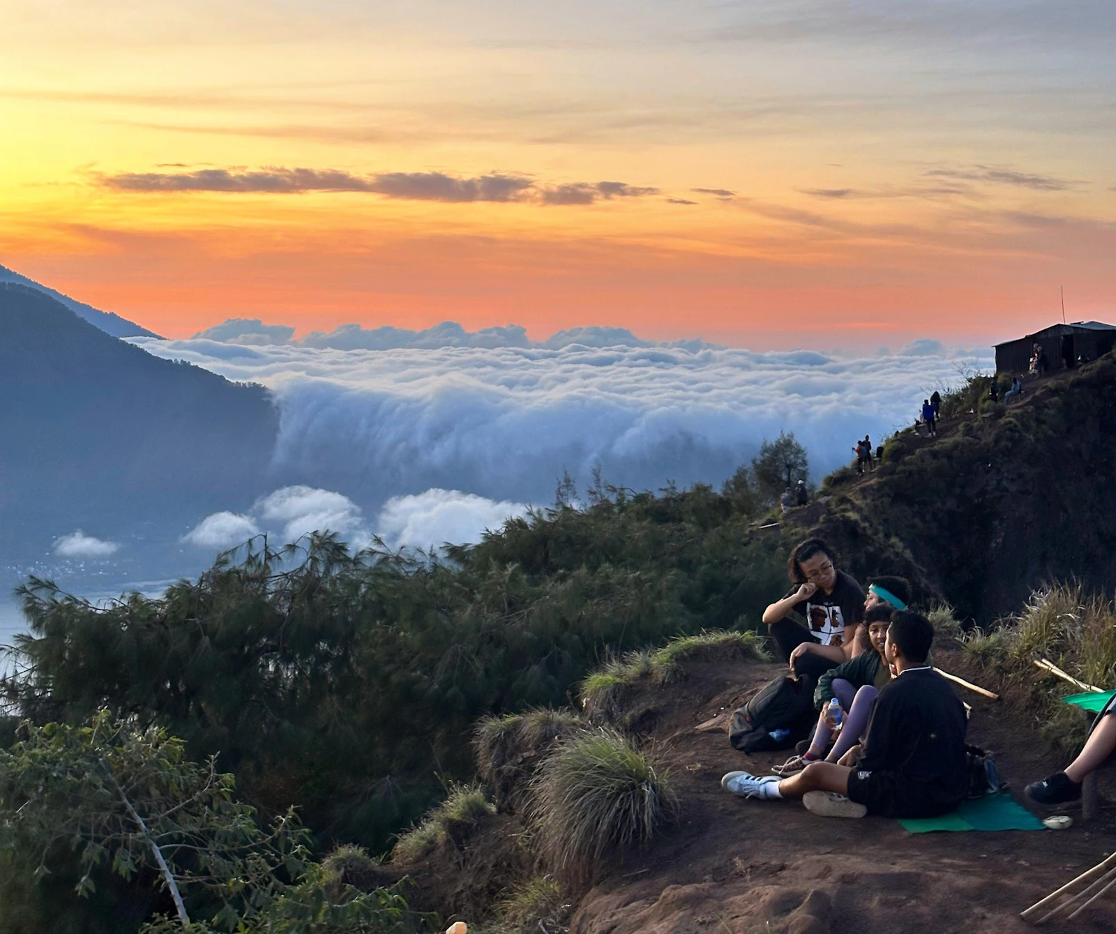 Hikers sit on a ridge above a sea of cloud as an orange sunrise glows on the horizon at Mount Batur, Bali