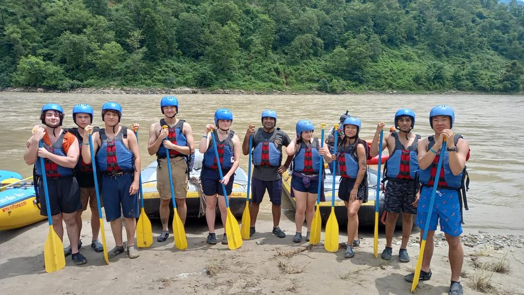 A line of students in helmets and buoyancy aids hold paddles beside a raft on a river bank.