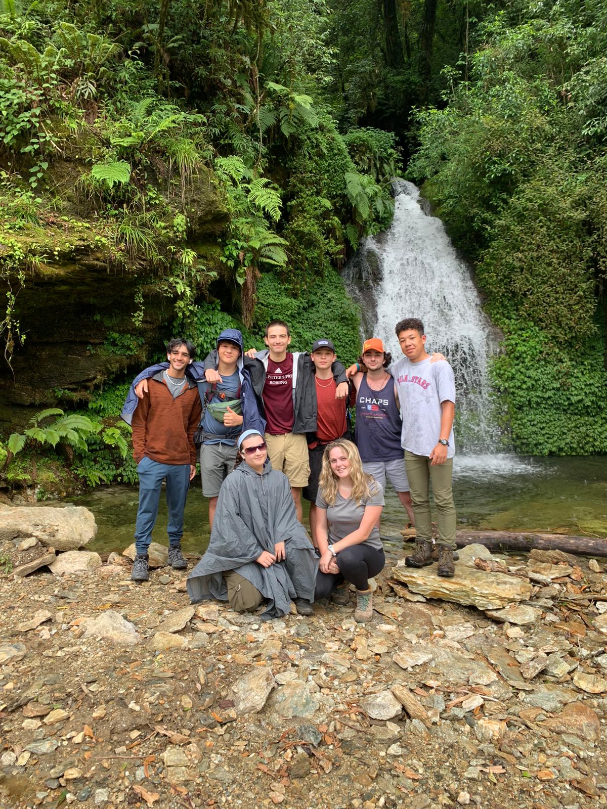 A group of students stands on rocks at the foot of a waterfall in dense green forest.