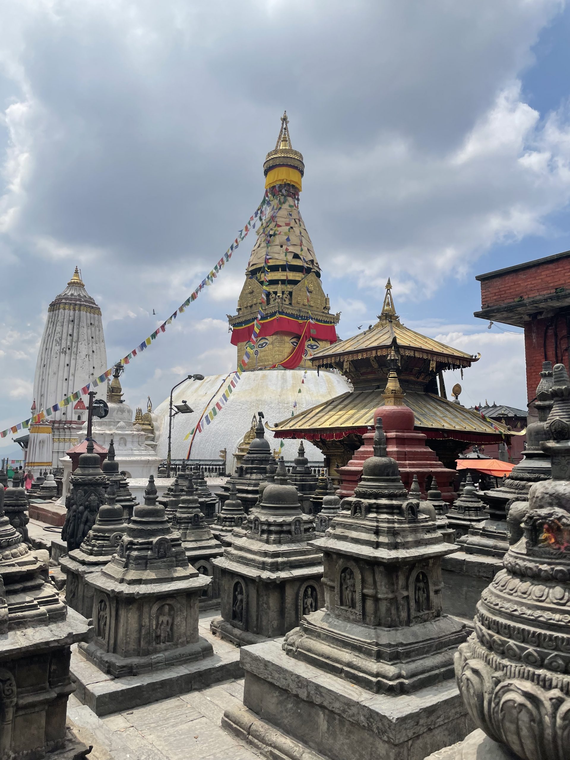 A whitewashed stupa with a gilded spire rises behind a courtyard of smaller stone shrines under strings of prayer flags.