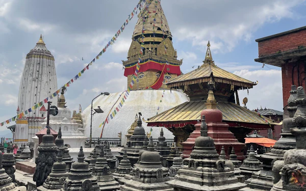 A whitewashed stupa with a gilded spire rises behind a courtyard of smaller stone shrines under strings of prayer flags.
