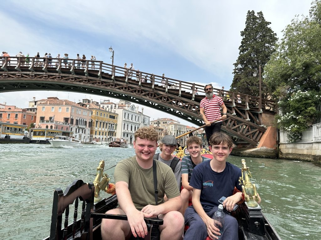Four students smile on a gondola ride in Venice, passing under a wooden bridge with colorful Venetian buildings in the background--one of the best things to do in Italy.
