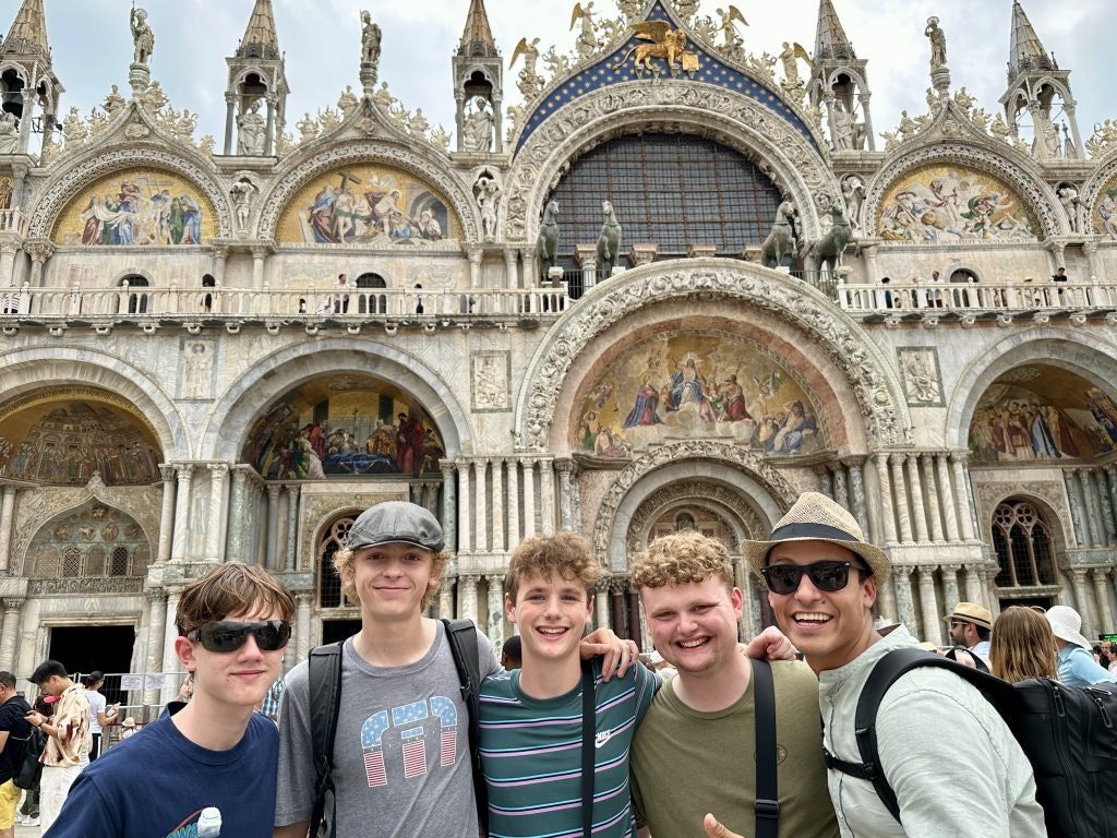 A teacher and students smiling together in front of St. Mark’s Basilica in Venice during a school trip, illustrating how free teacher travel supports meaningful experiences abroad.