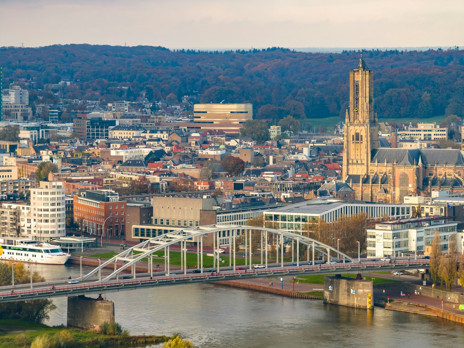 The white steel arch of the John Frost bridge over the Rhine at Arnhem, with the town, its church tower and a wooded ridge in autumn colour behind.