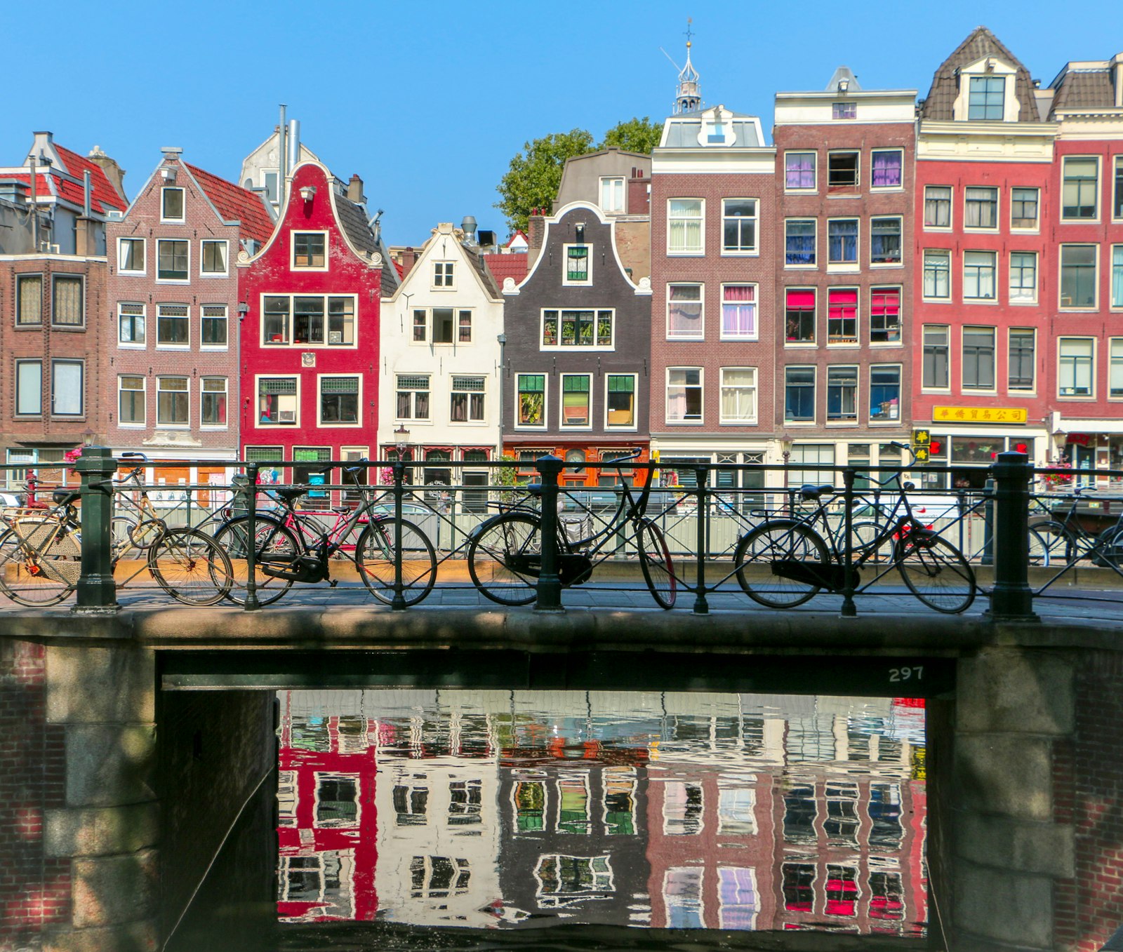 Bicycles leaning along a canal bridge railing in Amsterdam, a terrace of red, cream and grey gabled houses behind them and reflected in the water under the arch.