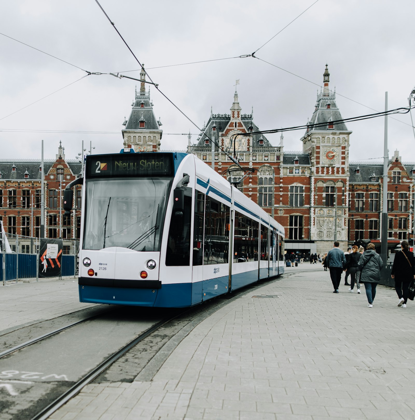 A blue and white tram waiting outside Amsterdam Centraal station, the brick and stone facade and its clock tower behind.