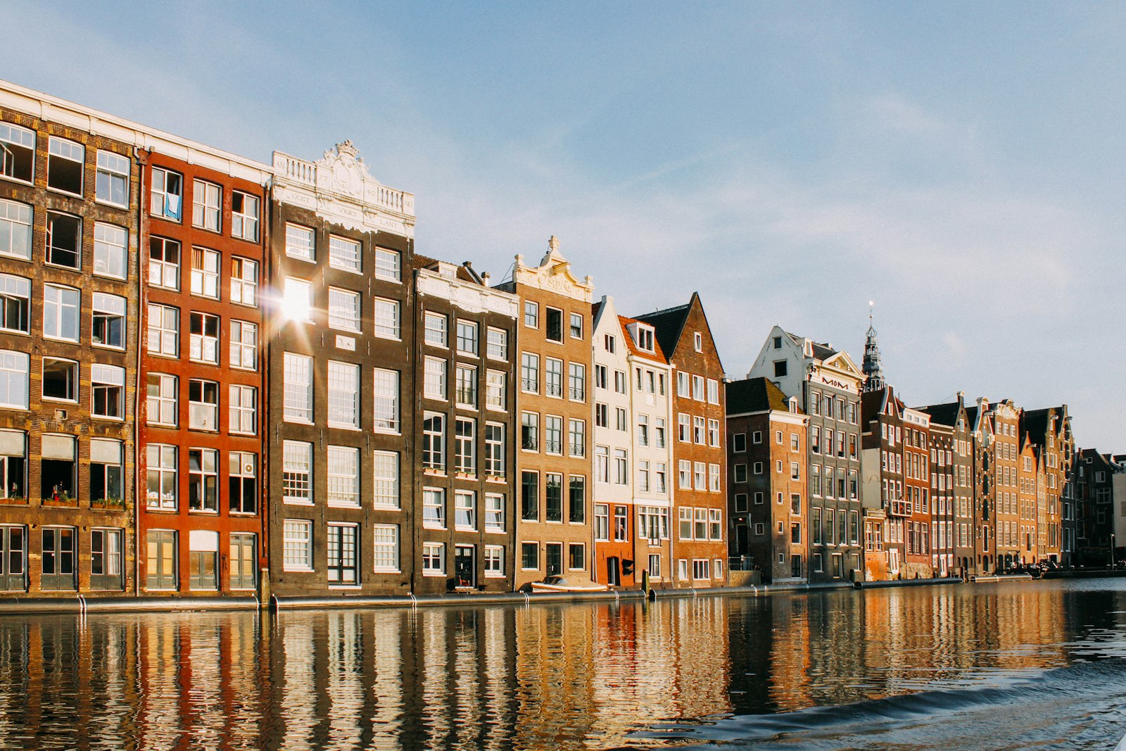 A row of narrow gabled canal houses in Amsterdam, low sun on the brick fronts and the whole row reflected in still water.