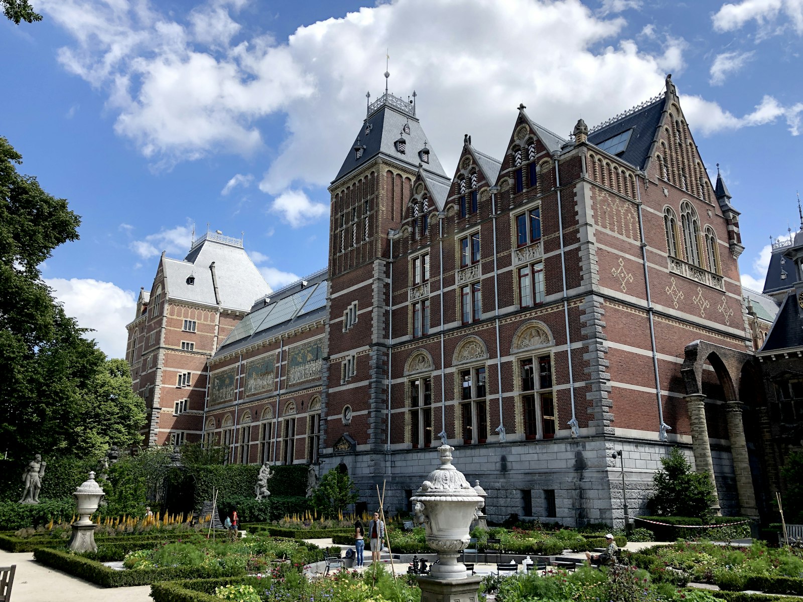 The Rijksmuseum seen across its formal garden, gabled brick and stone towers under a cumulus sky, with clipped hedges and stone urns in front.