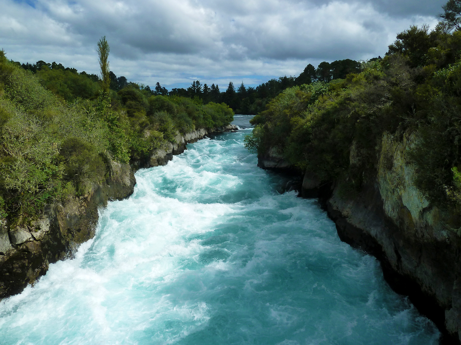 Turquoise river rushes through a forested gorge under cloudy skies
