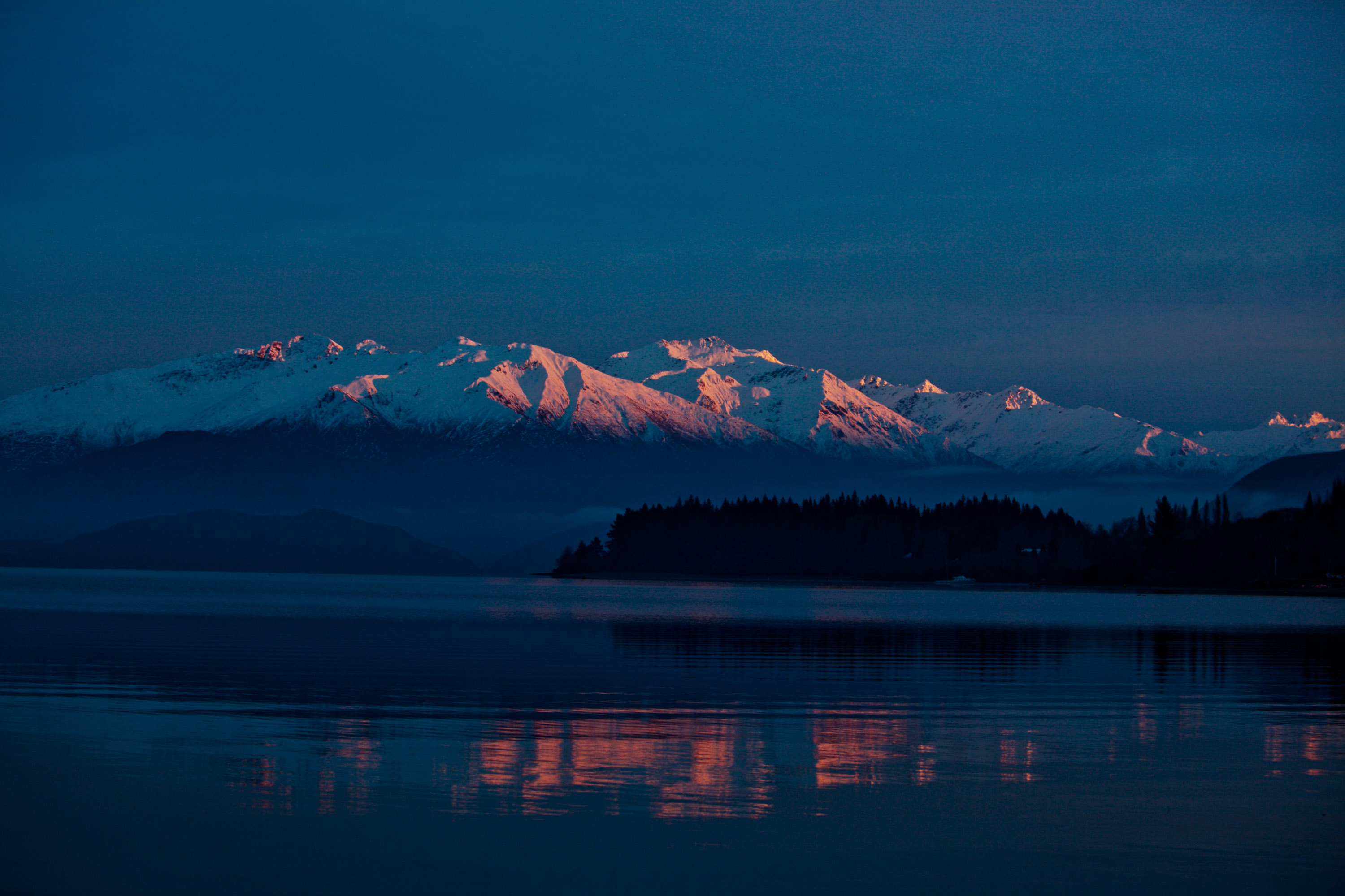 A snow-covered range catching the last pink light above a dark forested shore and still water.