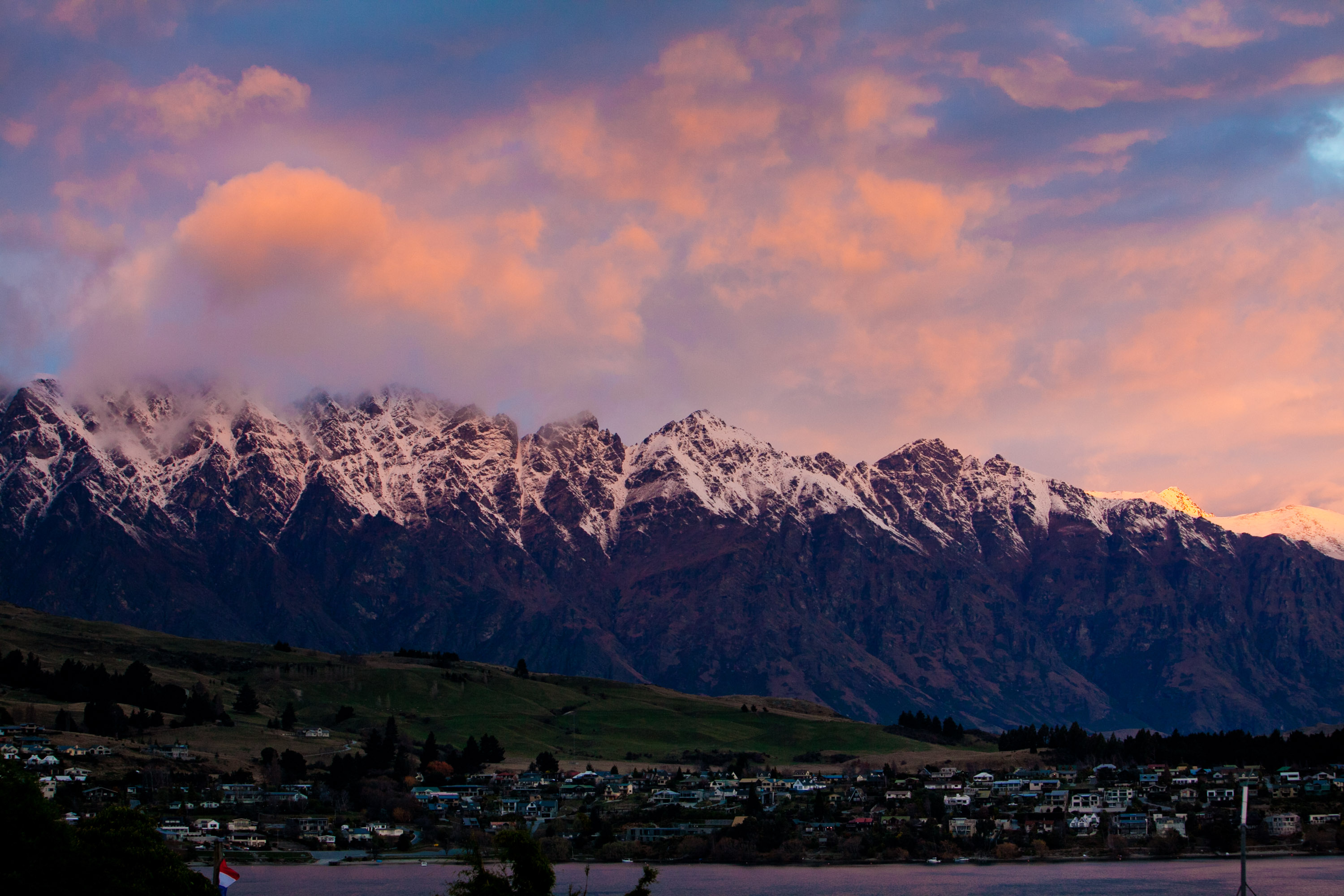 A jagged snow-dusted range lit pink at sunset above a lakeside town and its hillsides.