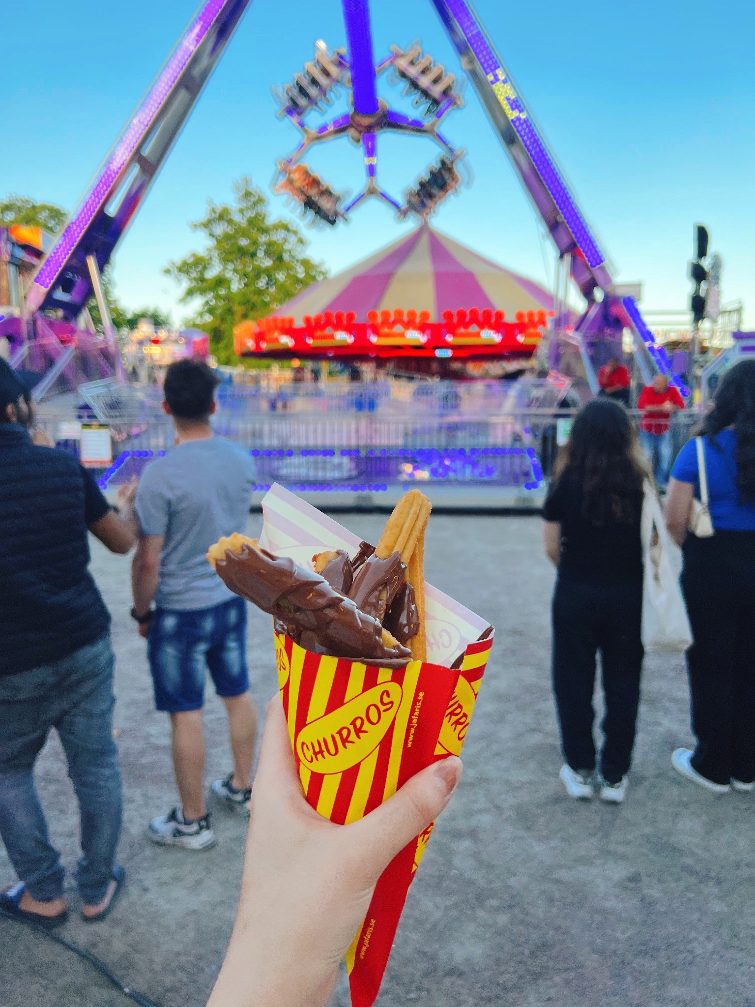 A hand holds a cone of chocolate-covered churros at an amusement park with a swing ride and striped tent in the background