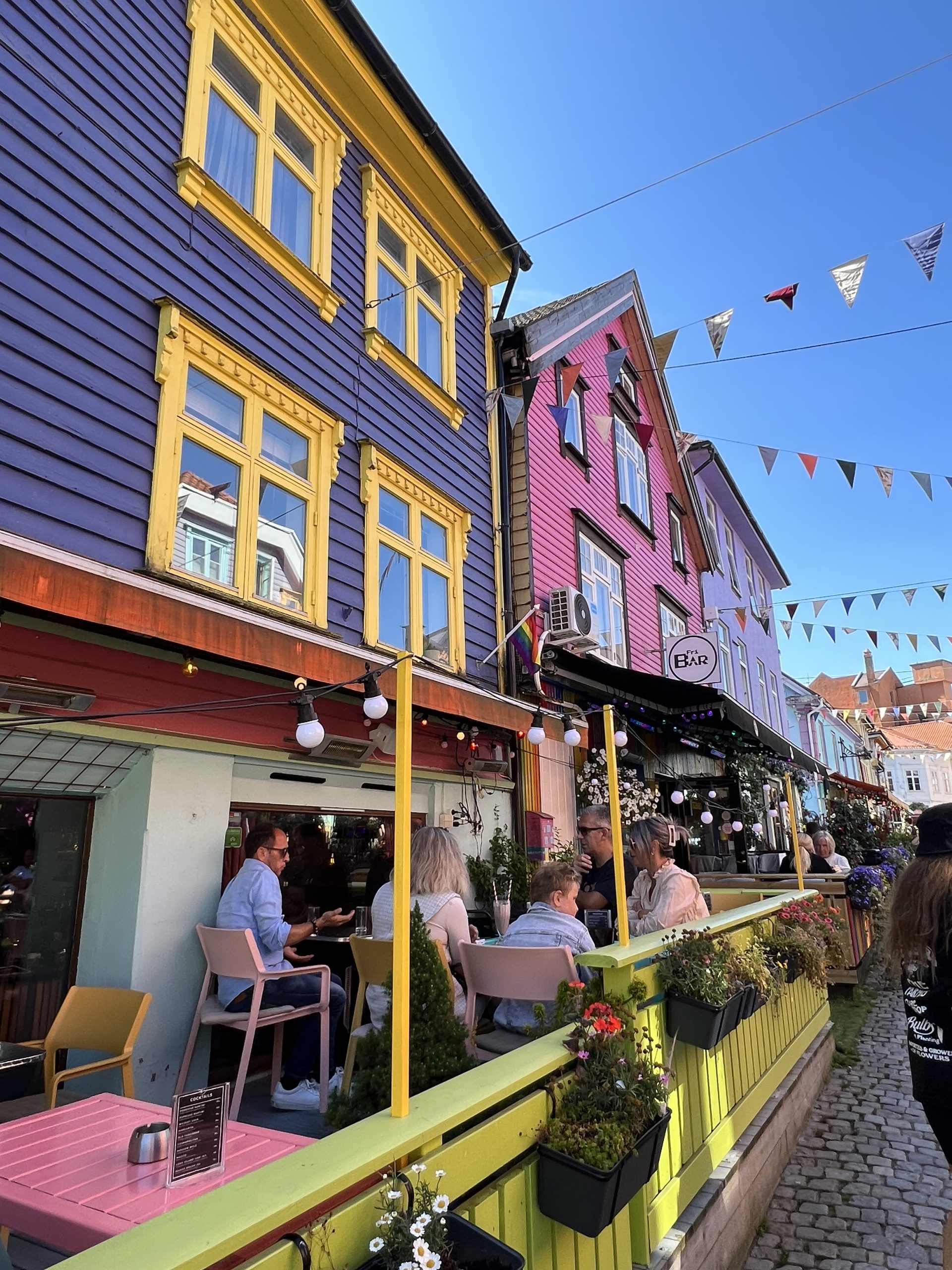 Diners sit at a café along a cobblestone street lined with colorful wooden buildings and bunting