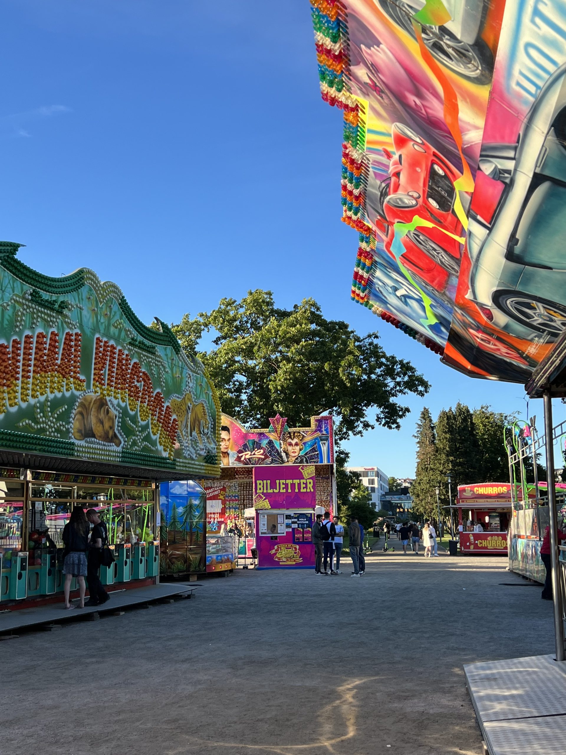 Colorful ride banners and a ticket booth line a fairground midway under blue sky