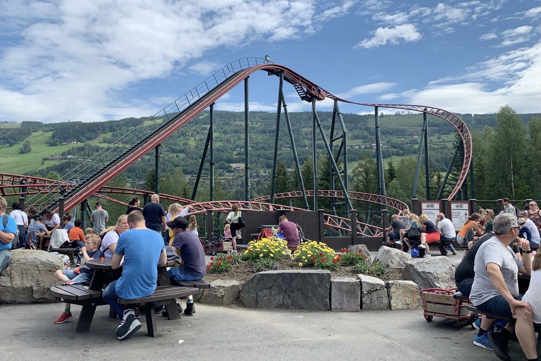 Visitors relax at picnic tables near a roller coaster winding through a green valley