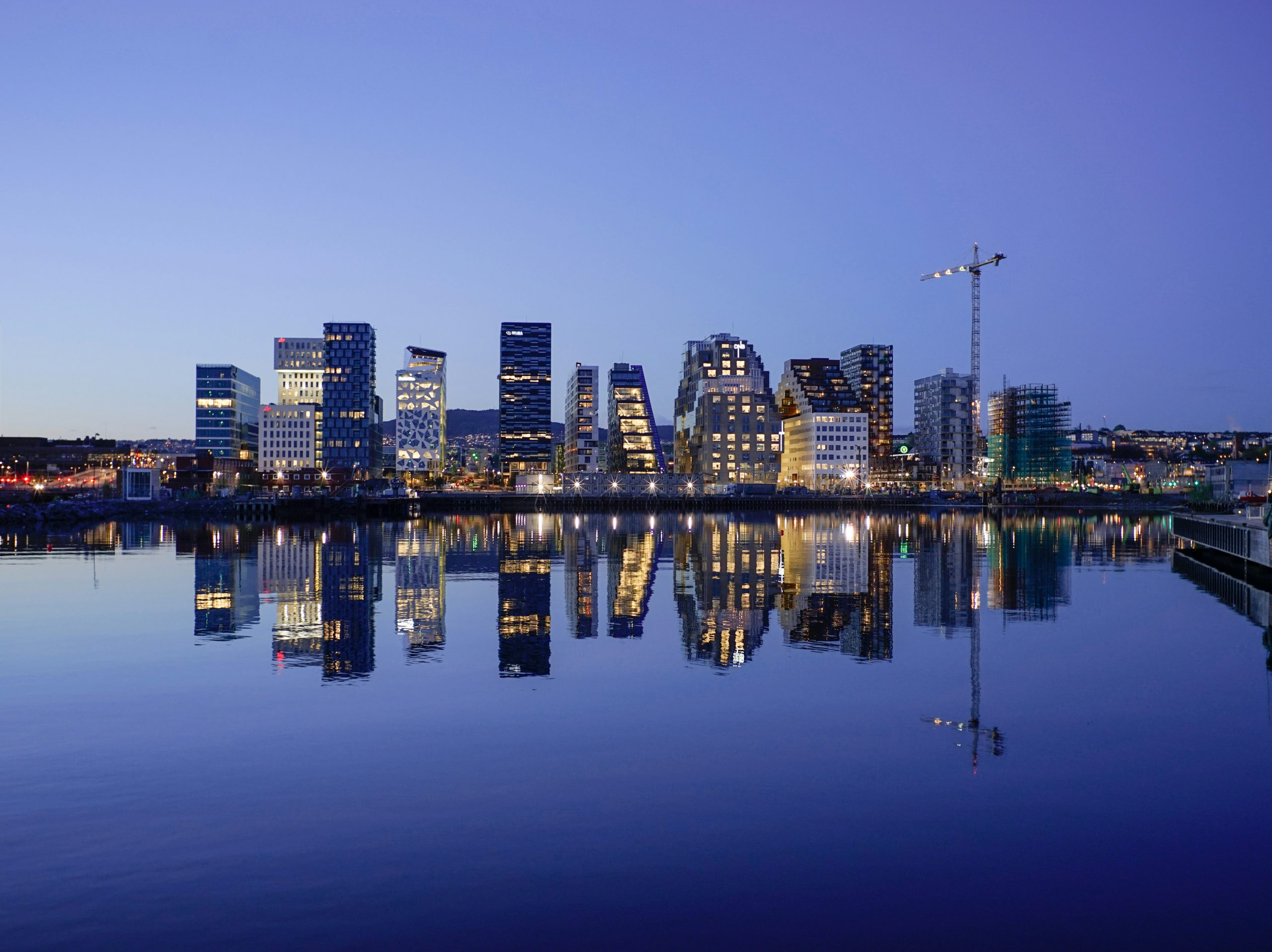 The modern high-rise buildings of Oslo's waterfront district glow at dusk, reflected in the harbor water.