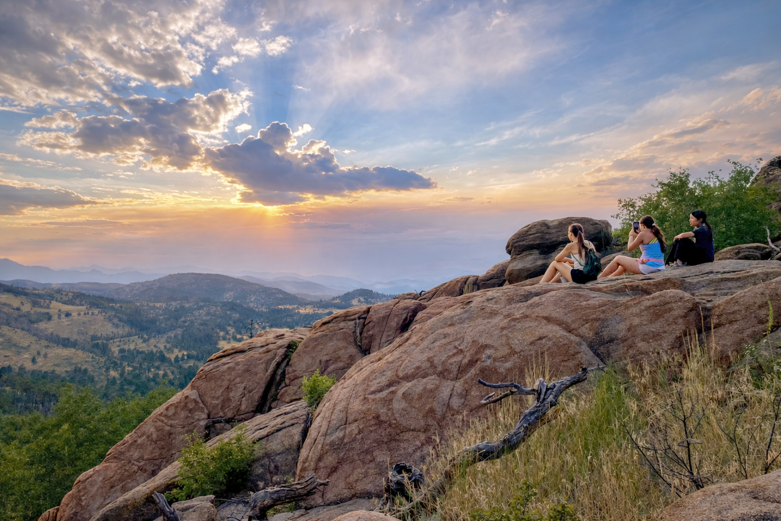Teen travelers sit on a rocky outcrop watching the sunset over mountain ridges