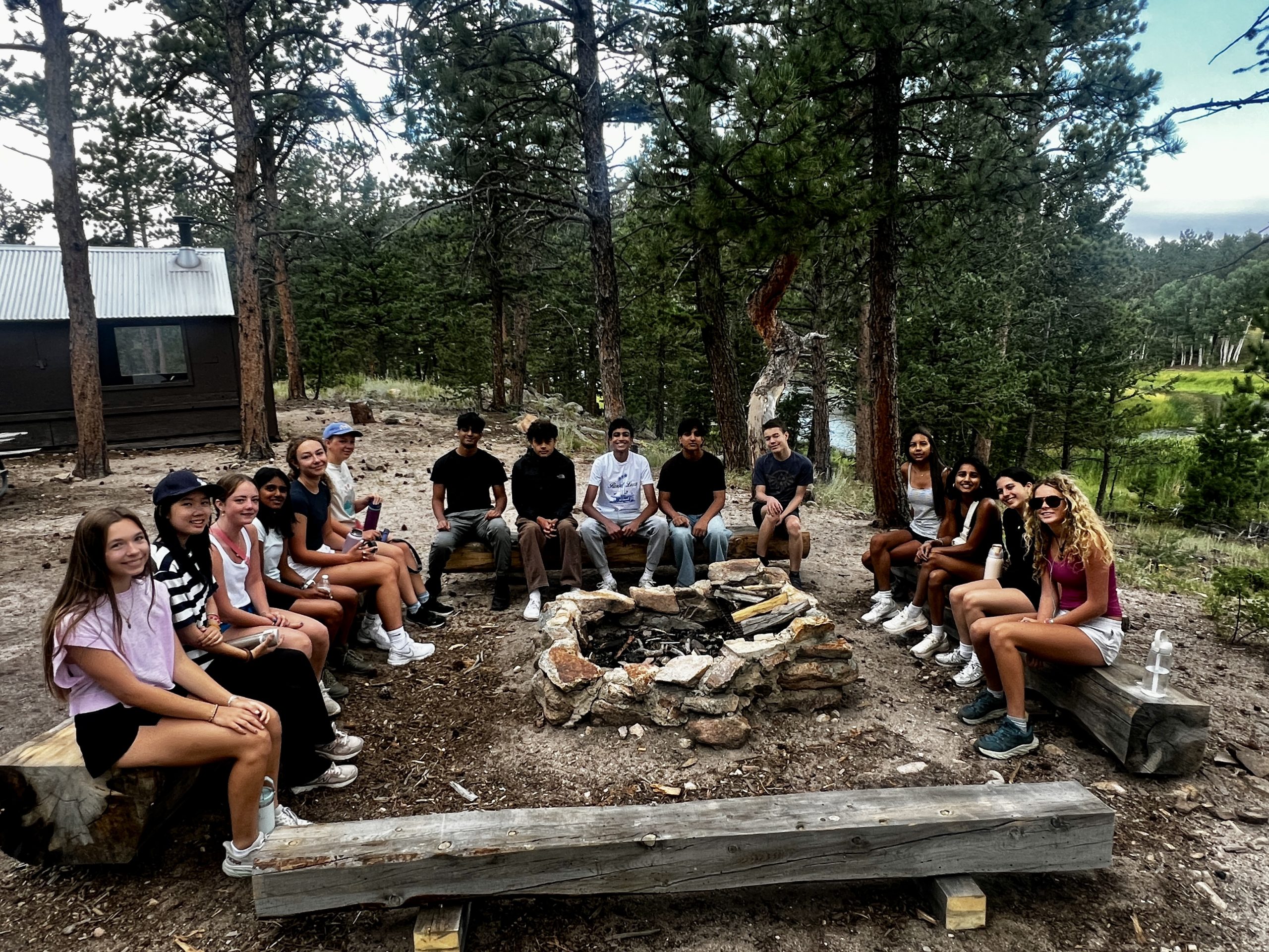 Group of teen campers sit around a stone fire pit at camp in the Colorado mountains after a day of activities.