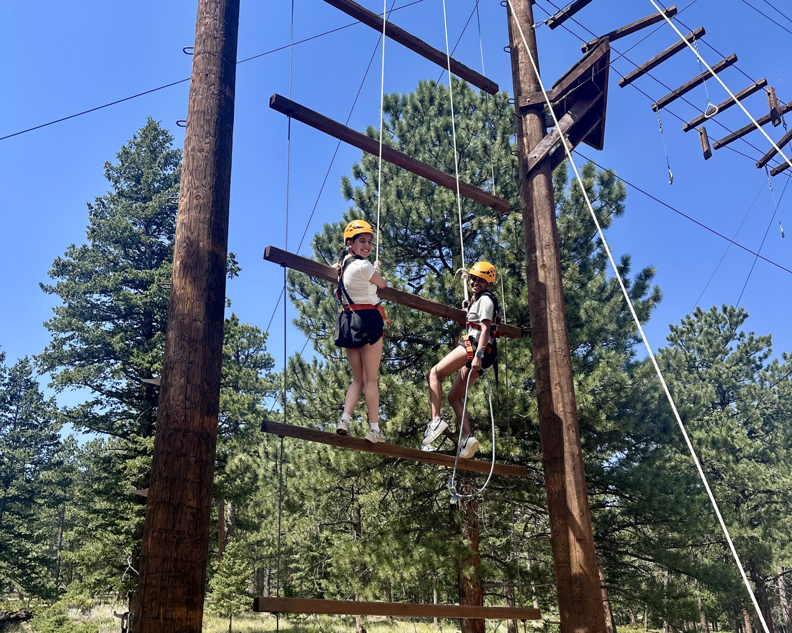 Two teen campers in helmets and harnesses climb a suspended ladder element on a Colorado high ropes course.