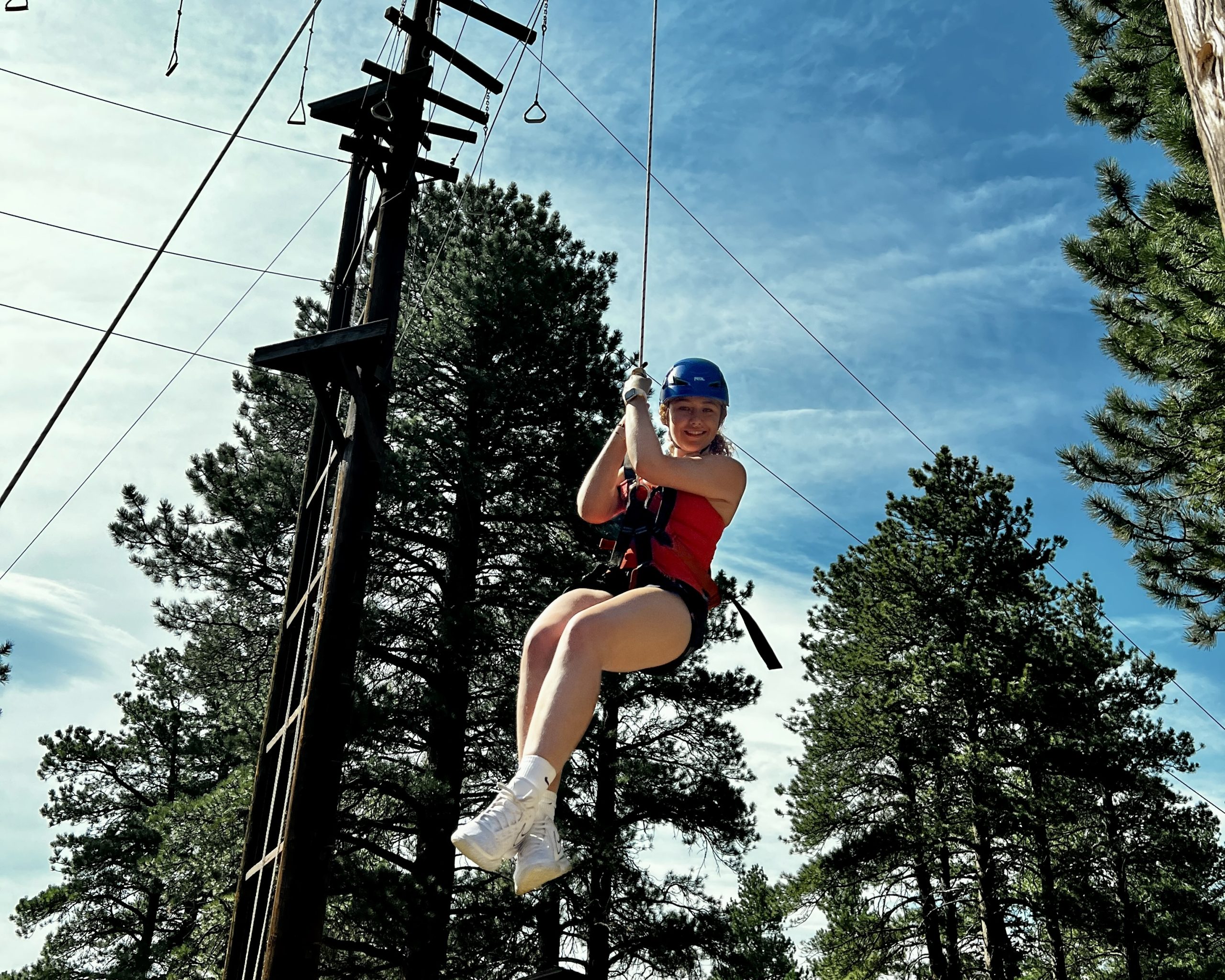 Teen rides a zip line through pine trees at a Colorado outdoor adventure camp ropes course.