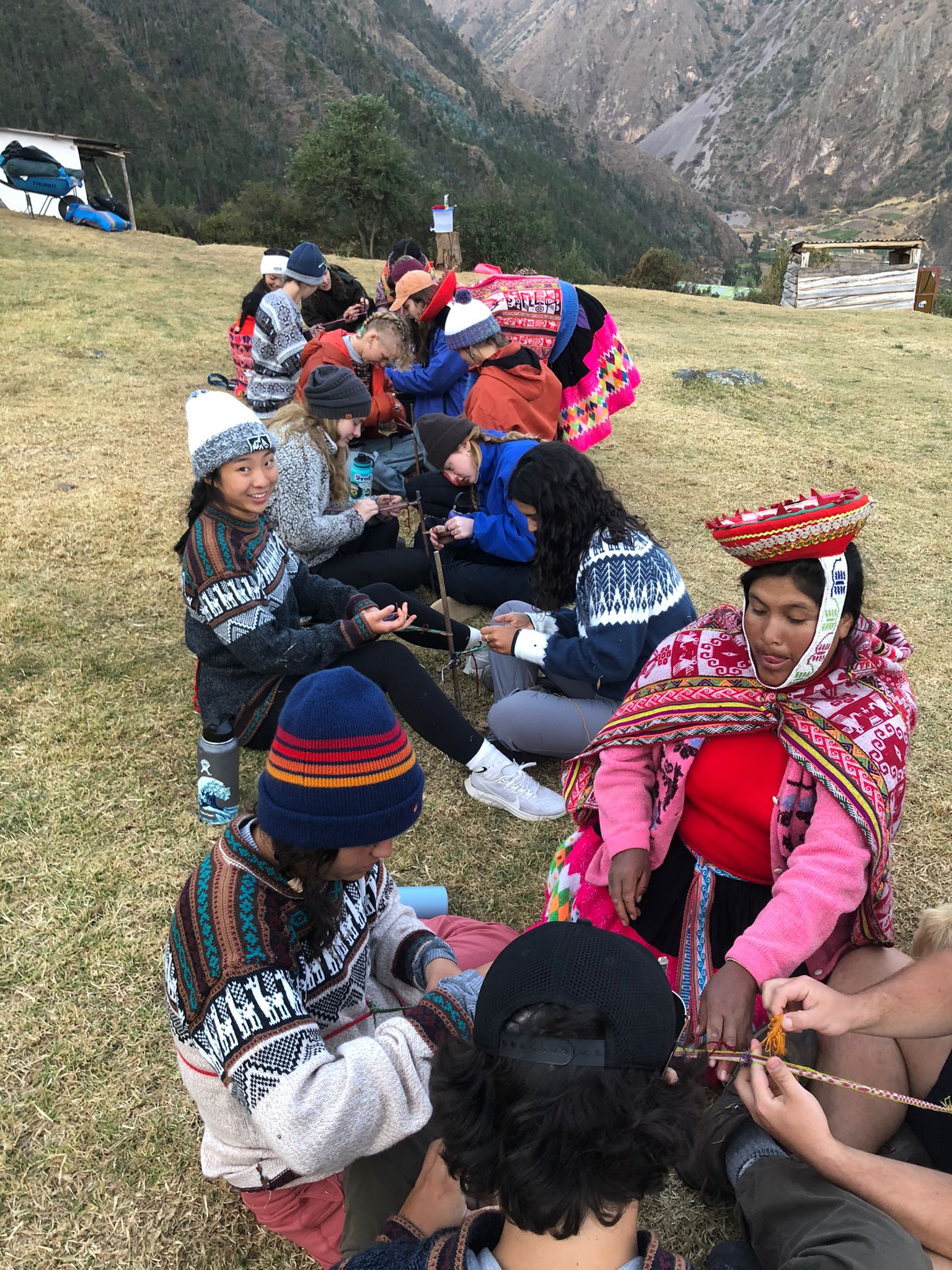 Students learn traditional weaving from a local artisan in colorful Andean dress on a grassy mountainside